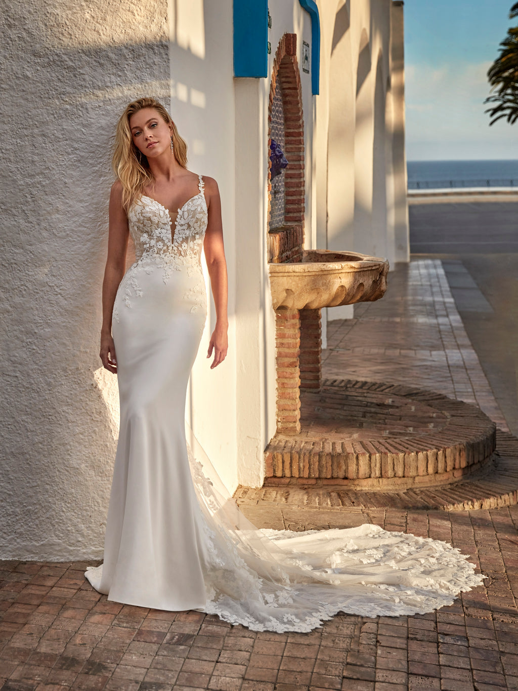 Woman in a white wedding dress with lace details standing outdoors near a fountain.