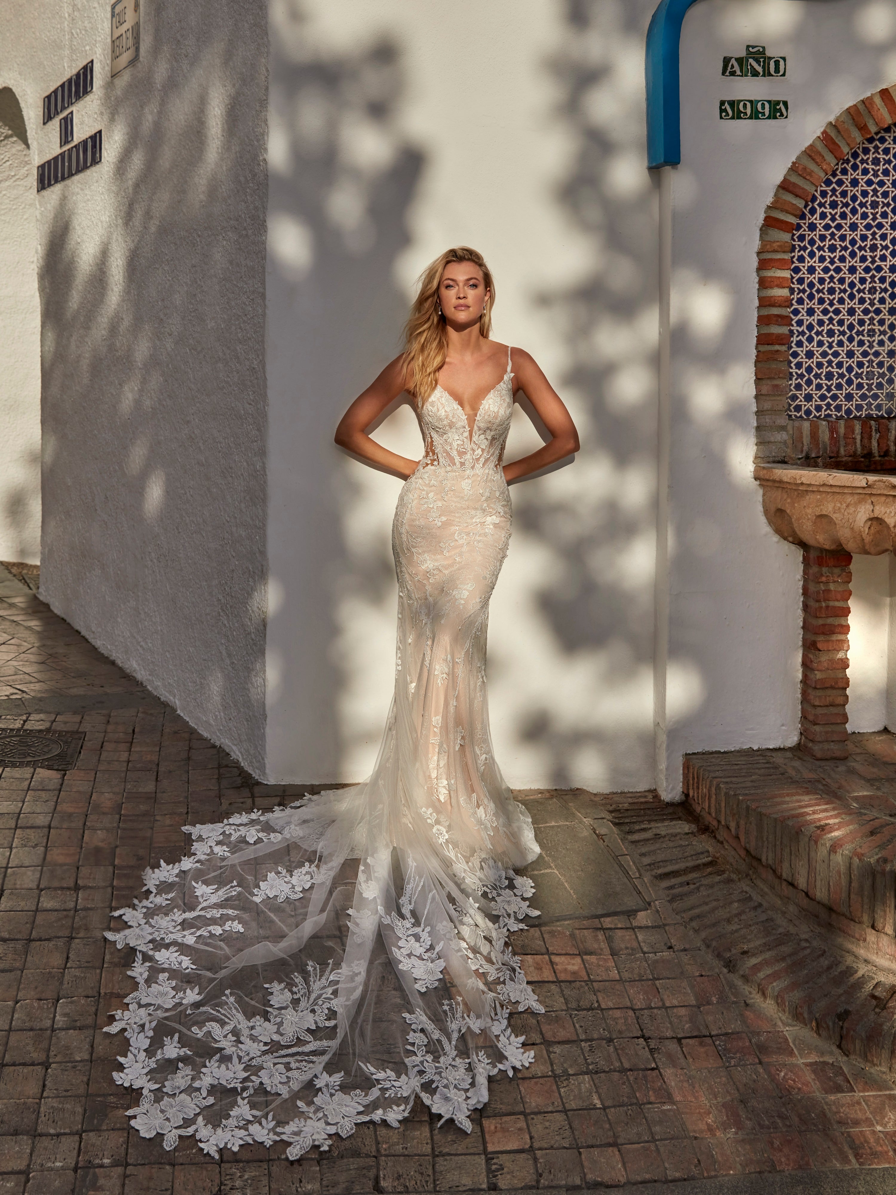 Woman in a white wedding dress with a long train standing in a sunlit outdoor setting.