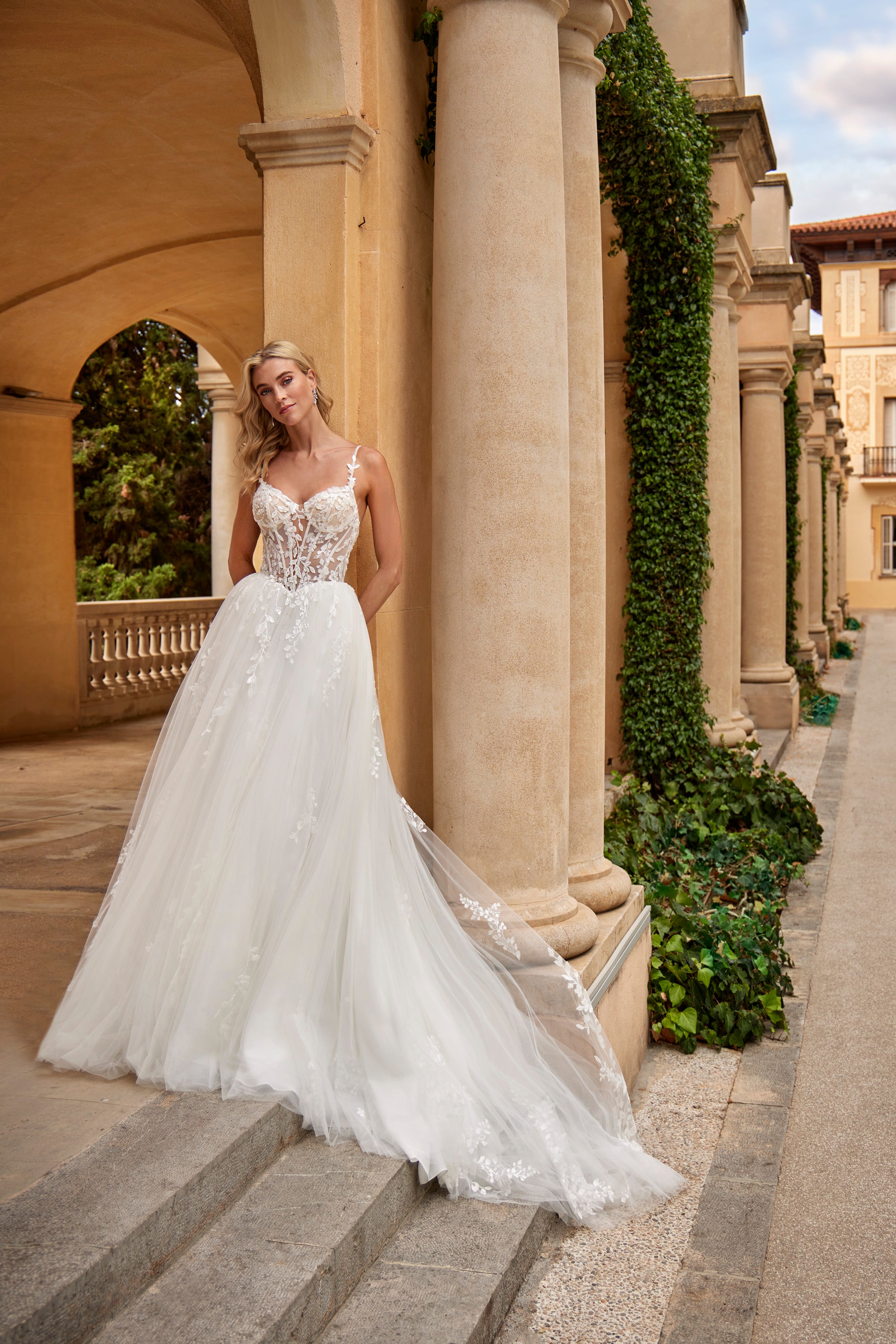 Woman in a white wedding dress standing in an elegant outdoor setting with columns and greenery.