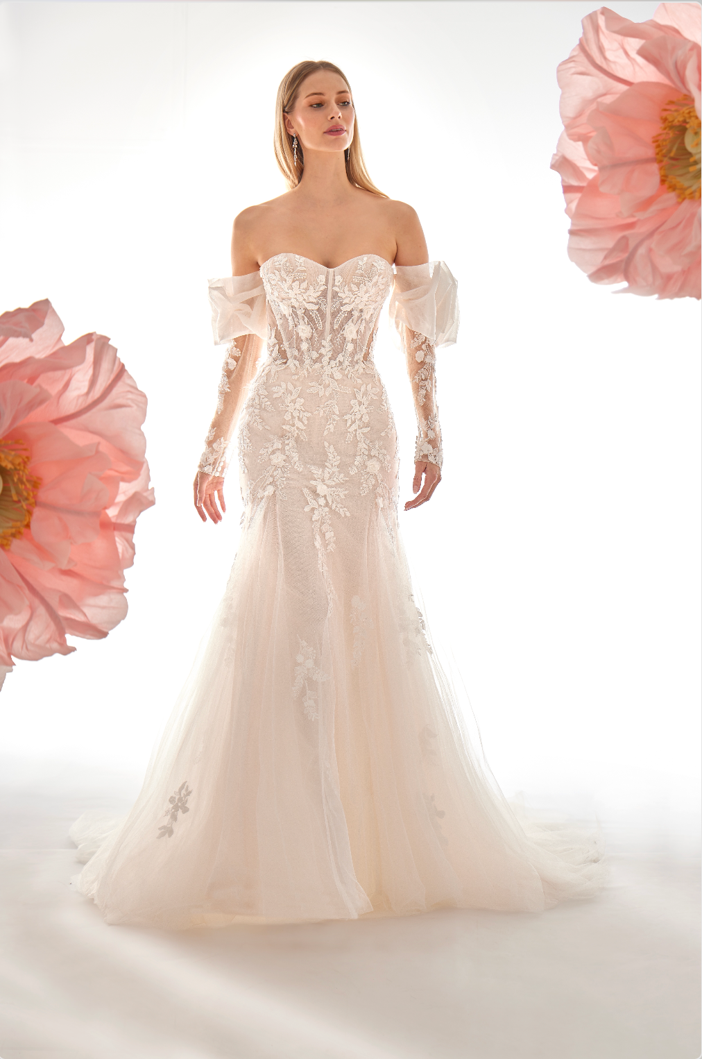 Woman wearing a white wedding dress with floral embellishments, surrounded by pink flowers on a light gray background.