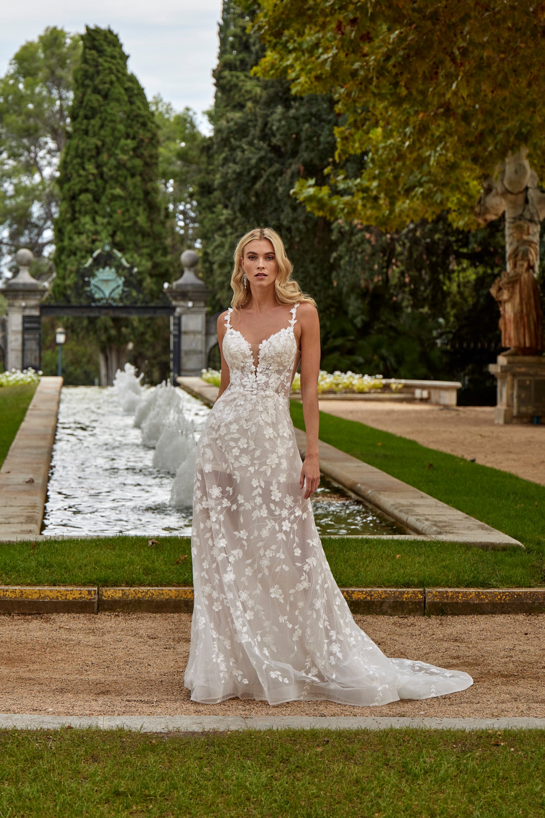 Woman in a floral wedding dress standing in a garden with a fountain.