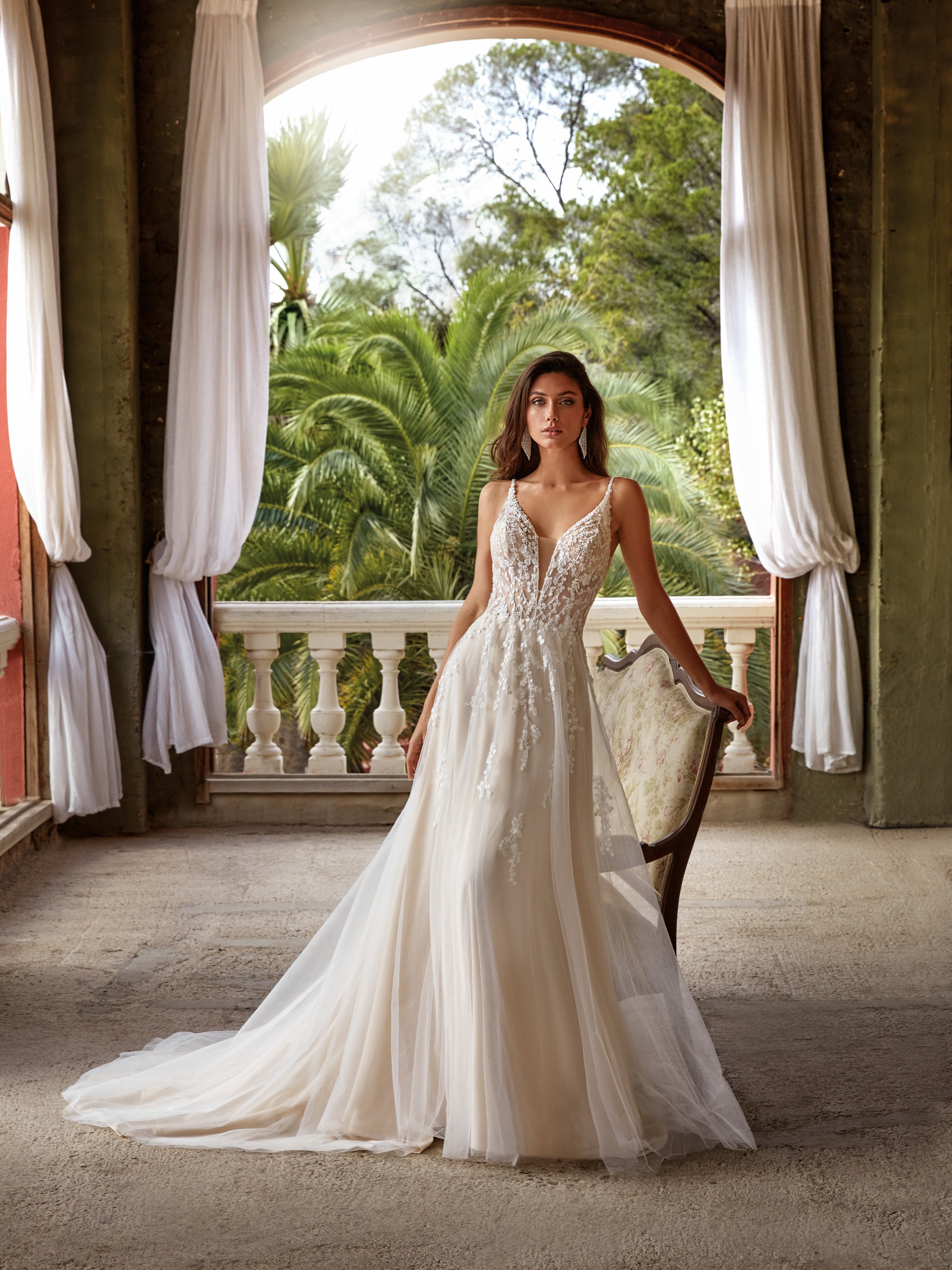 Woman in a white wedding dress standing in a grand hallway with greenery outside.