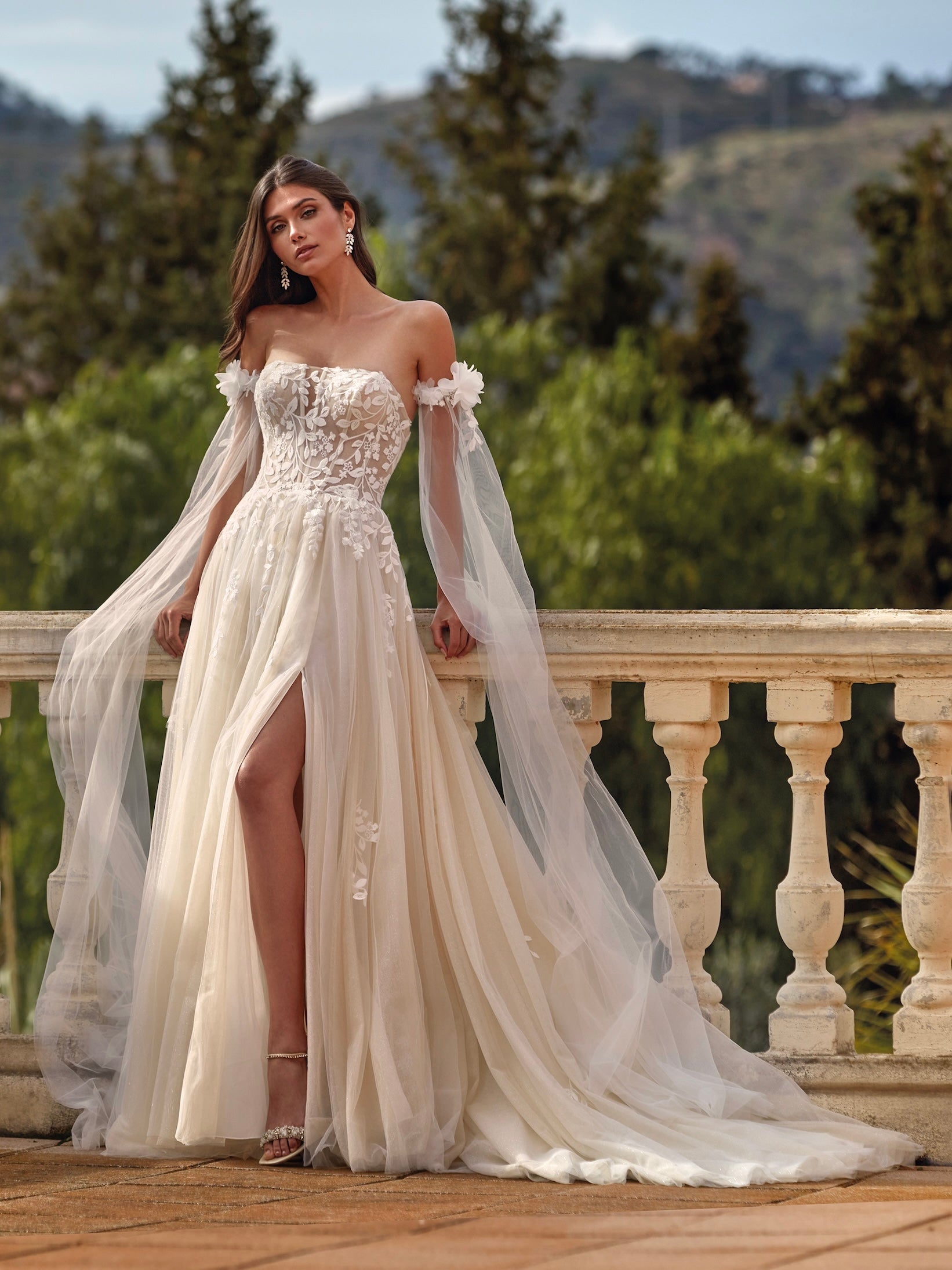 Woman in a white wedding dress with floral details standing on a balcony with greenery in the background
