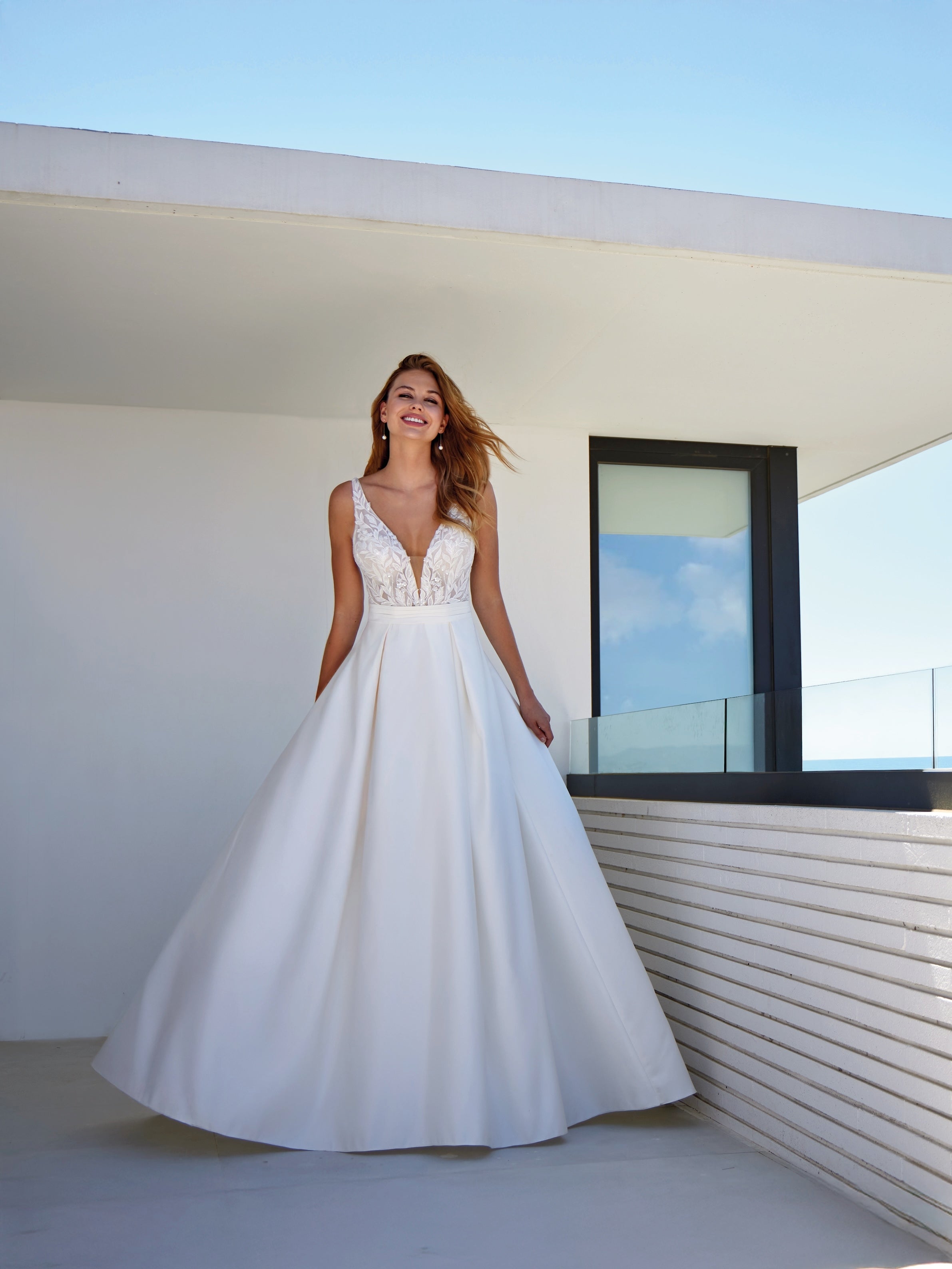 Woman in a white wedding dress standing on a balcony with a clear blue sky.