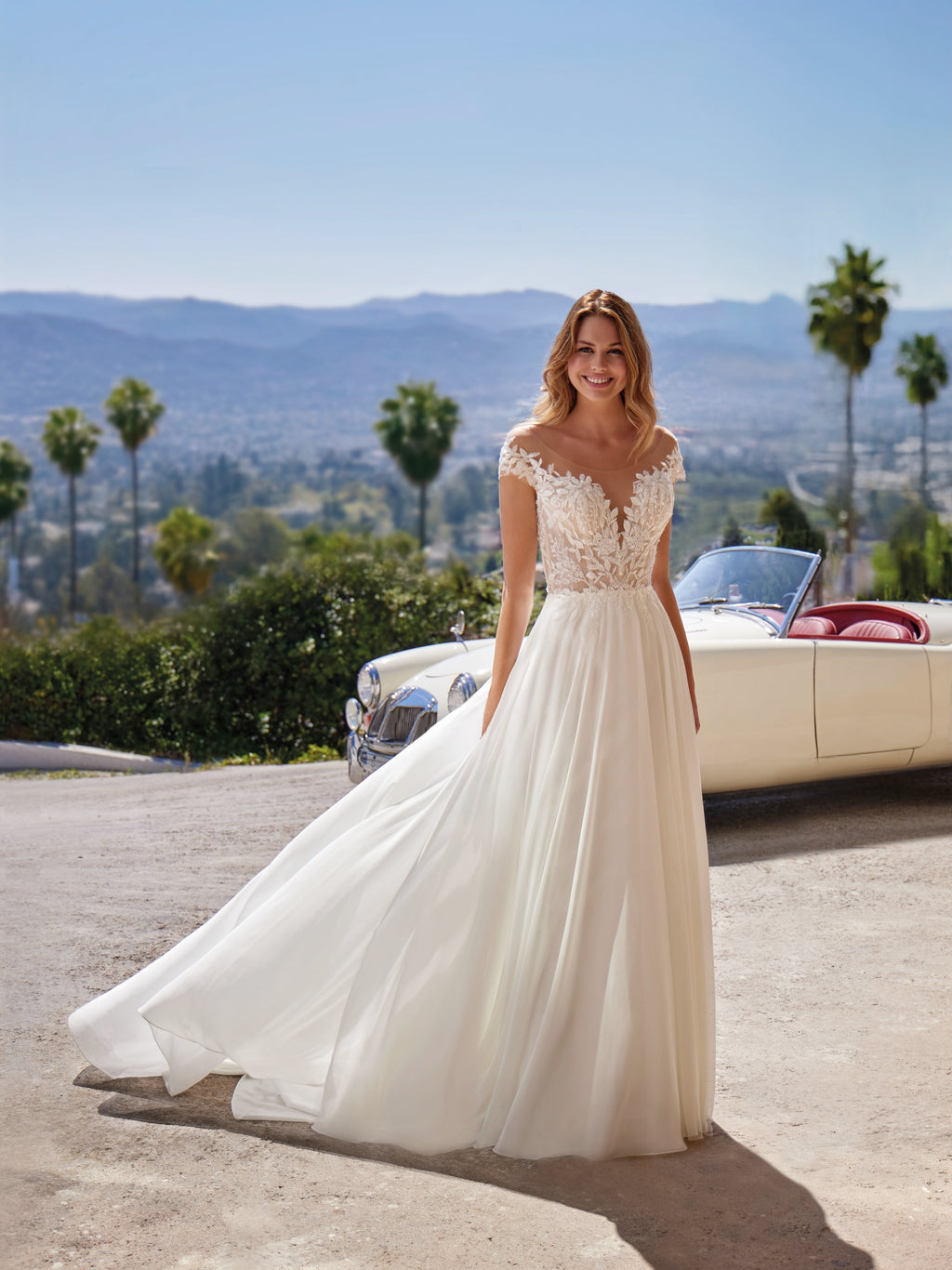 Woman in a white wedding dress standing in front of a vintage car with palm trees and mountains in the background.