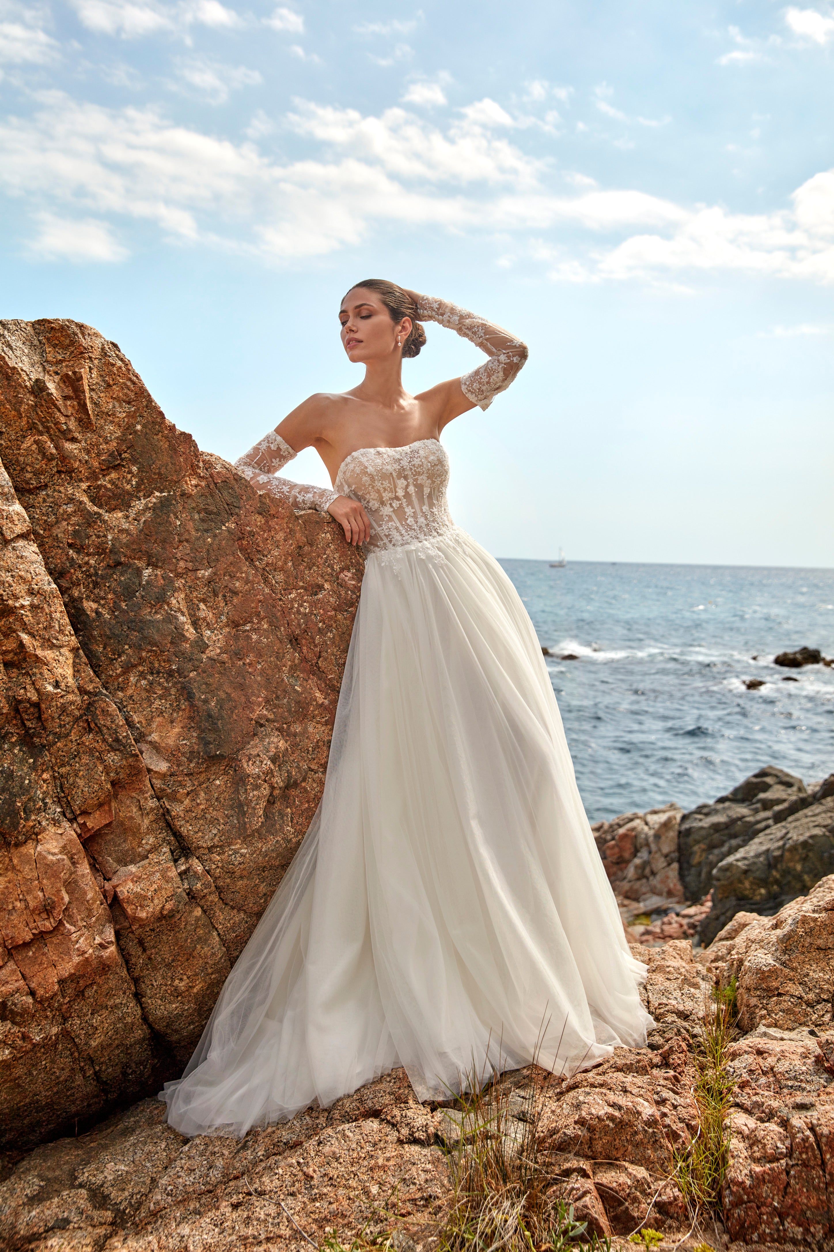 Woman in a white wedding dress standing on rocky terrain with ocean and sky in the background