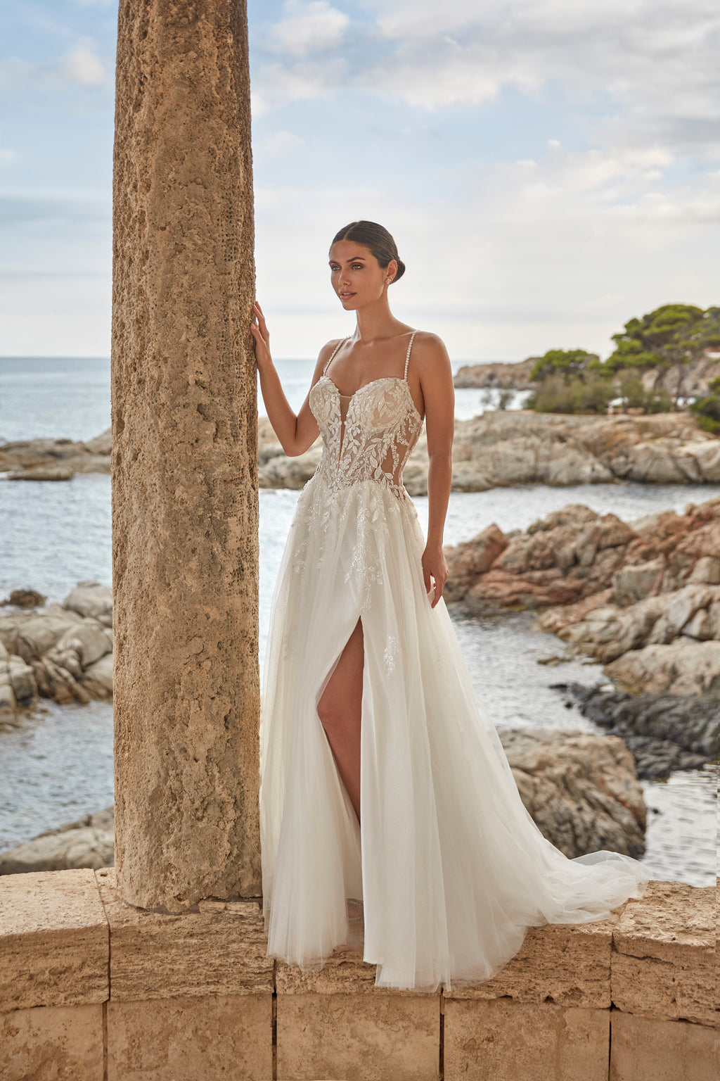 Woman in a white wedding dress standing by a stone column with ocean and rocks in the background