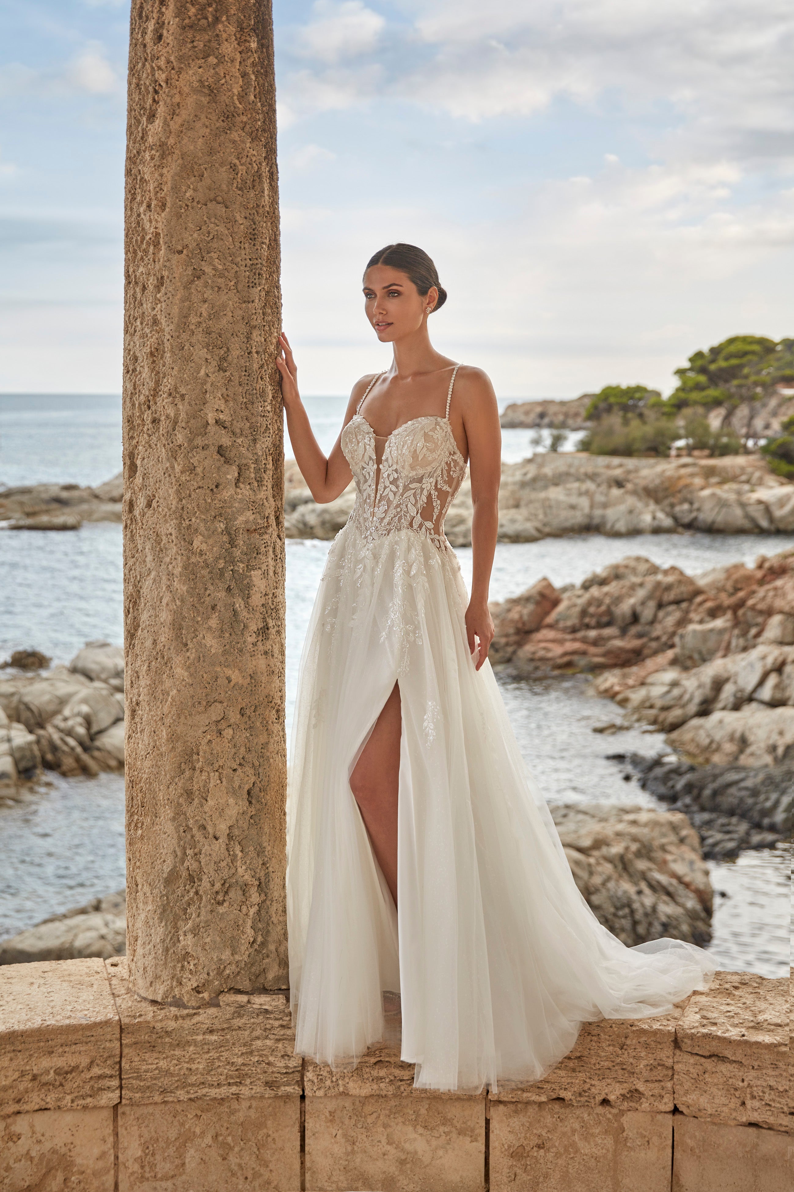 Woman in a white wedding dress standing by a stone column with ocean and rocks in the background