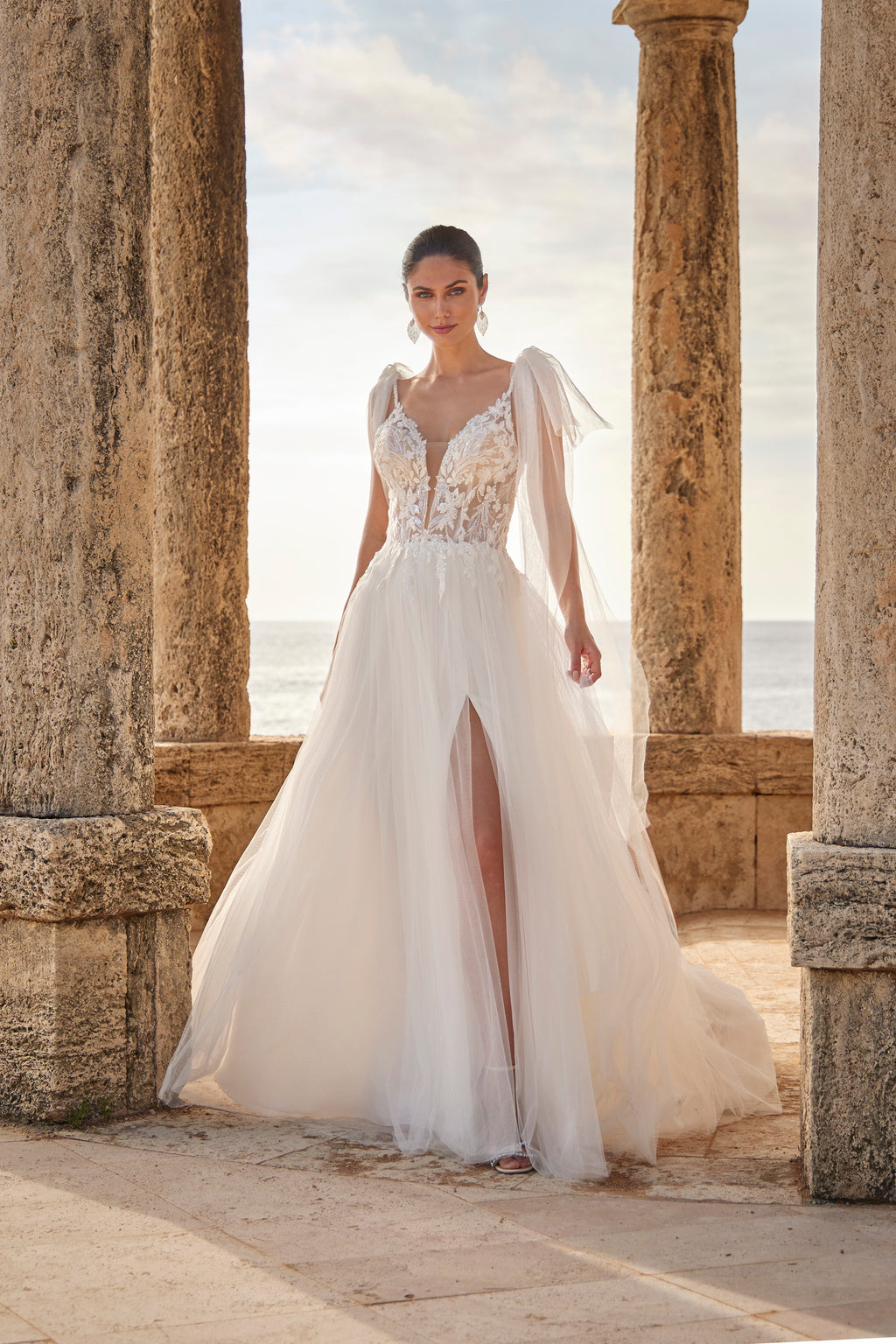 Woman in a white wedding dress with floral details standing in front of stone columns and ocean view.