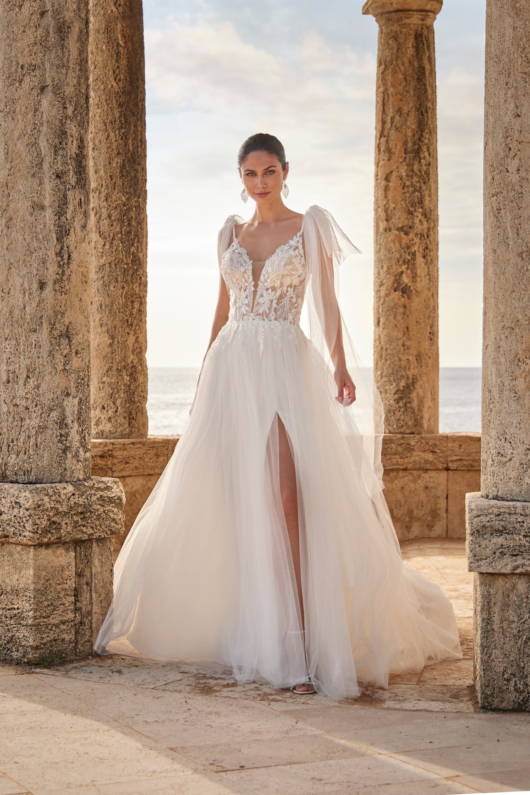 Woman in a white wedding dress with floral details standing in front of stone columns and ocean view.