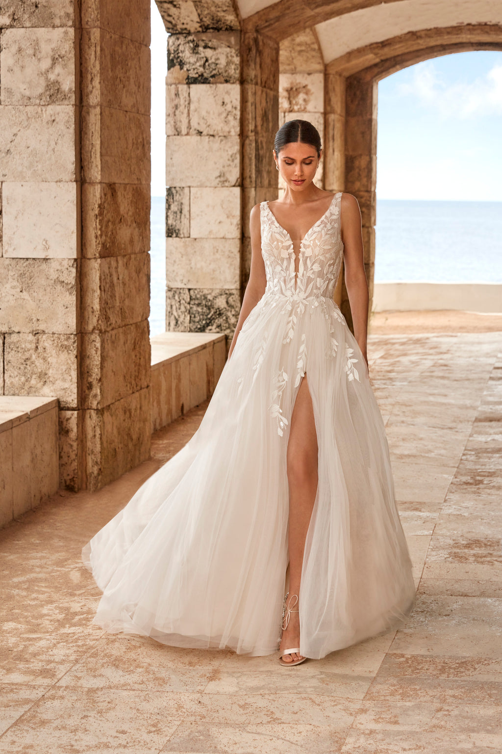 Woman in a white wedding dress standing in a stone archway with a scenic background.