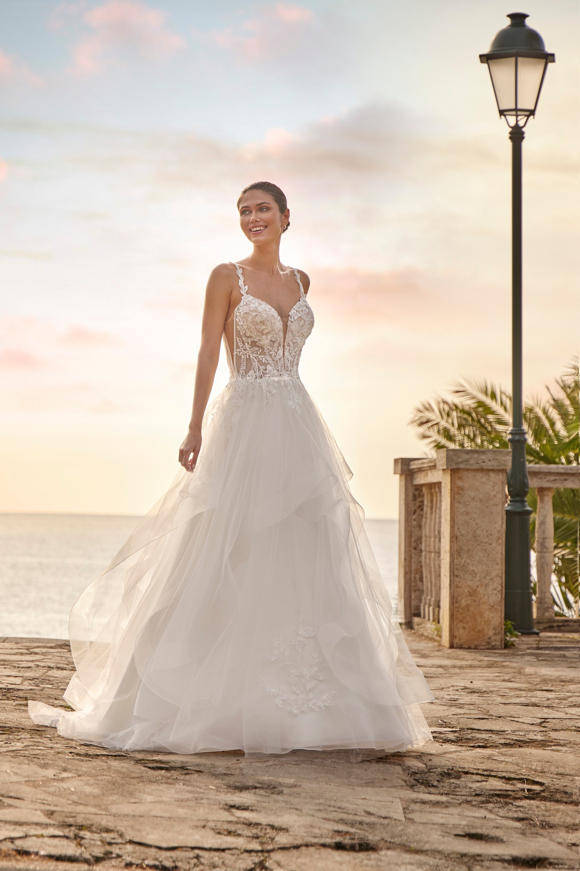 Woman in a white wedding dress standing on a beach with palm trees and a street lamp in the background.