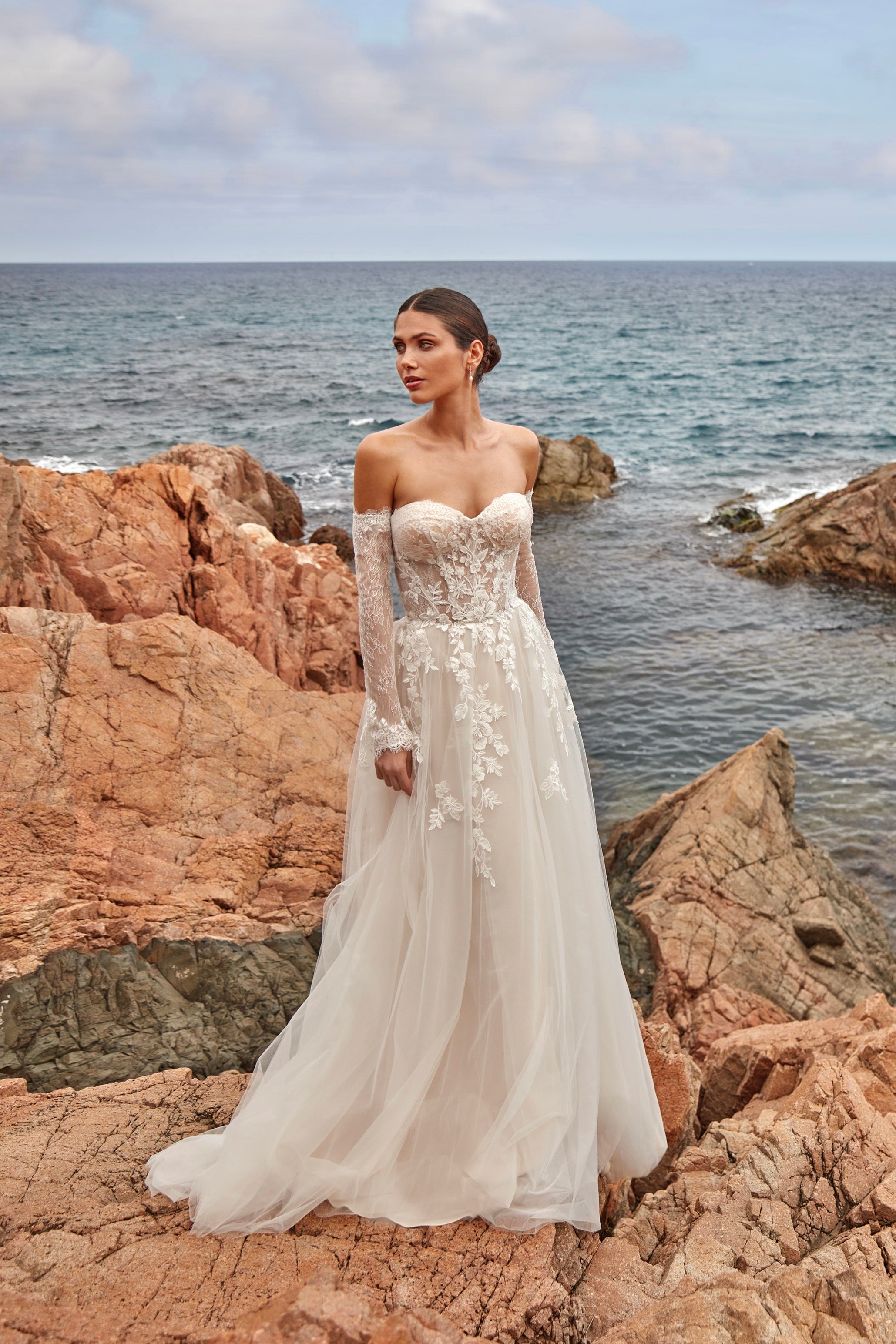 Woman in a white wedding dress standing on rocky coastal terrain with ocean in the background