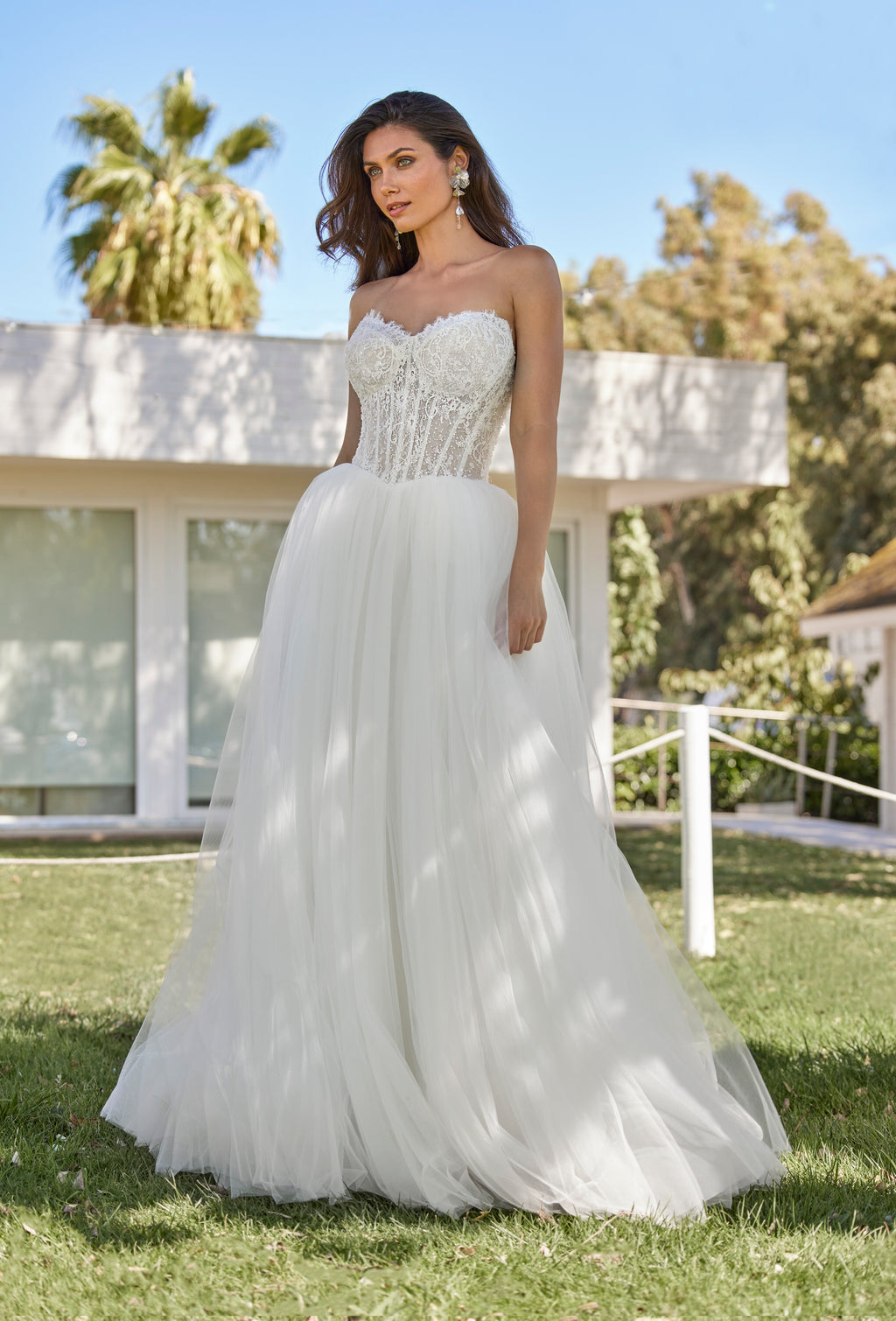 Woman in a white wedding dress standing outdoors with trees and a building in the background