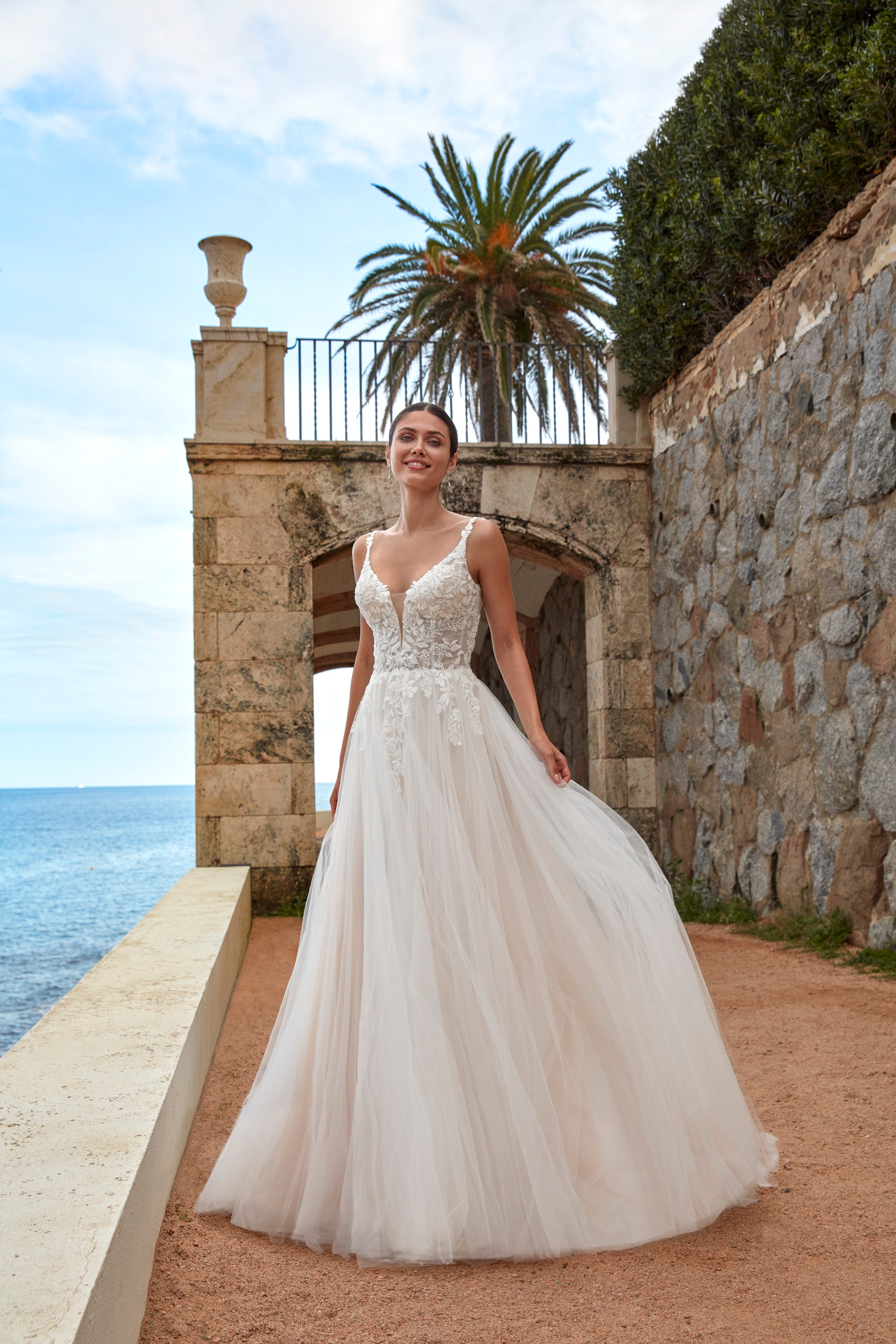 Woman in a white wedding dress standing in front of a stone wall and palm tree with ocean view.