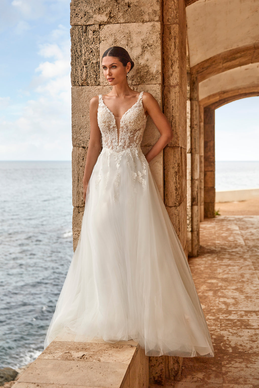 Woman in a white wedding dress standing by a stone wall with ocean view