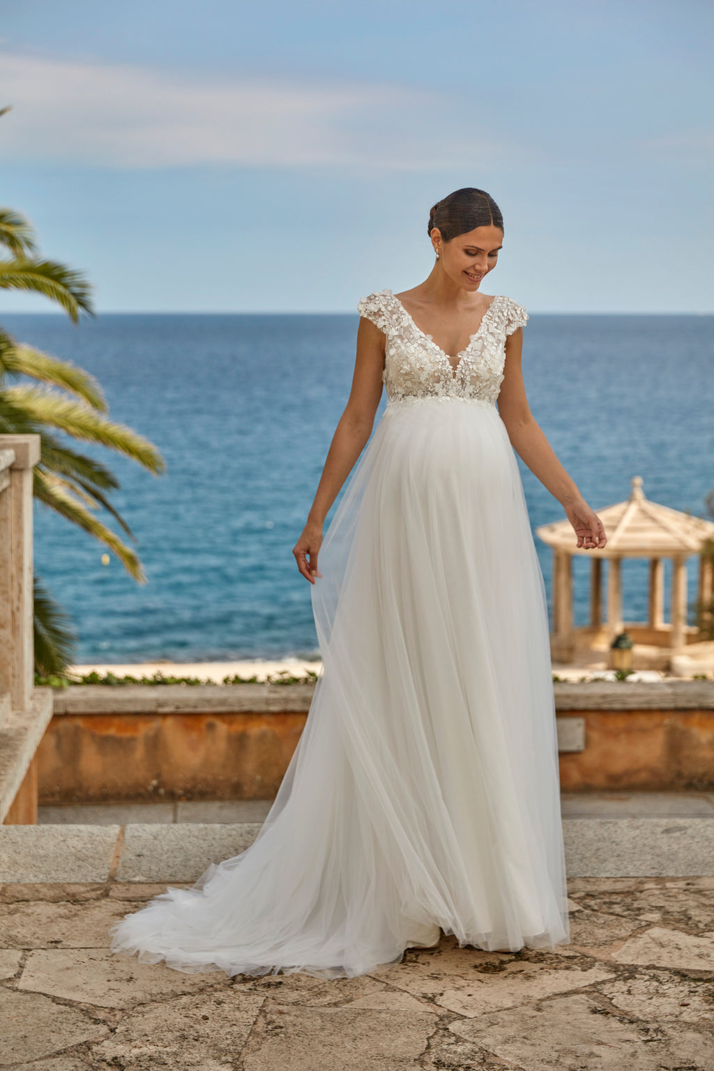 Woman in a white wedding dress standing by the ocean with a gazebo in the background.