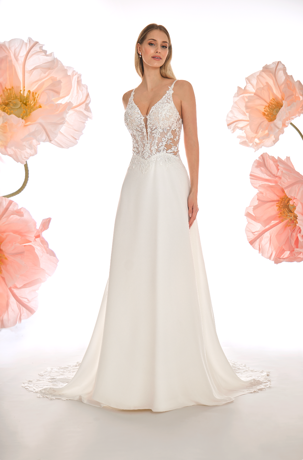 Woman wearing a white wedding dress with floral lace details, surrounded by pink flowers on a light background.
