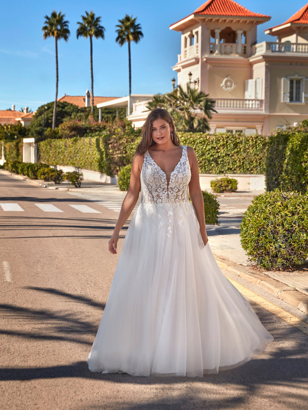 Woman in a white wedding dress standing on a street with palm trees and a building in the background.