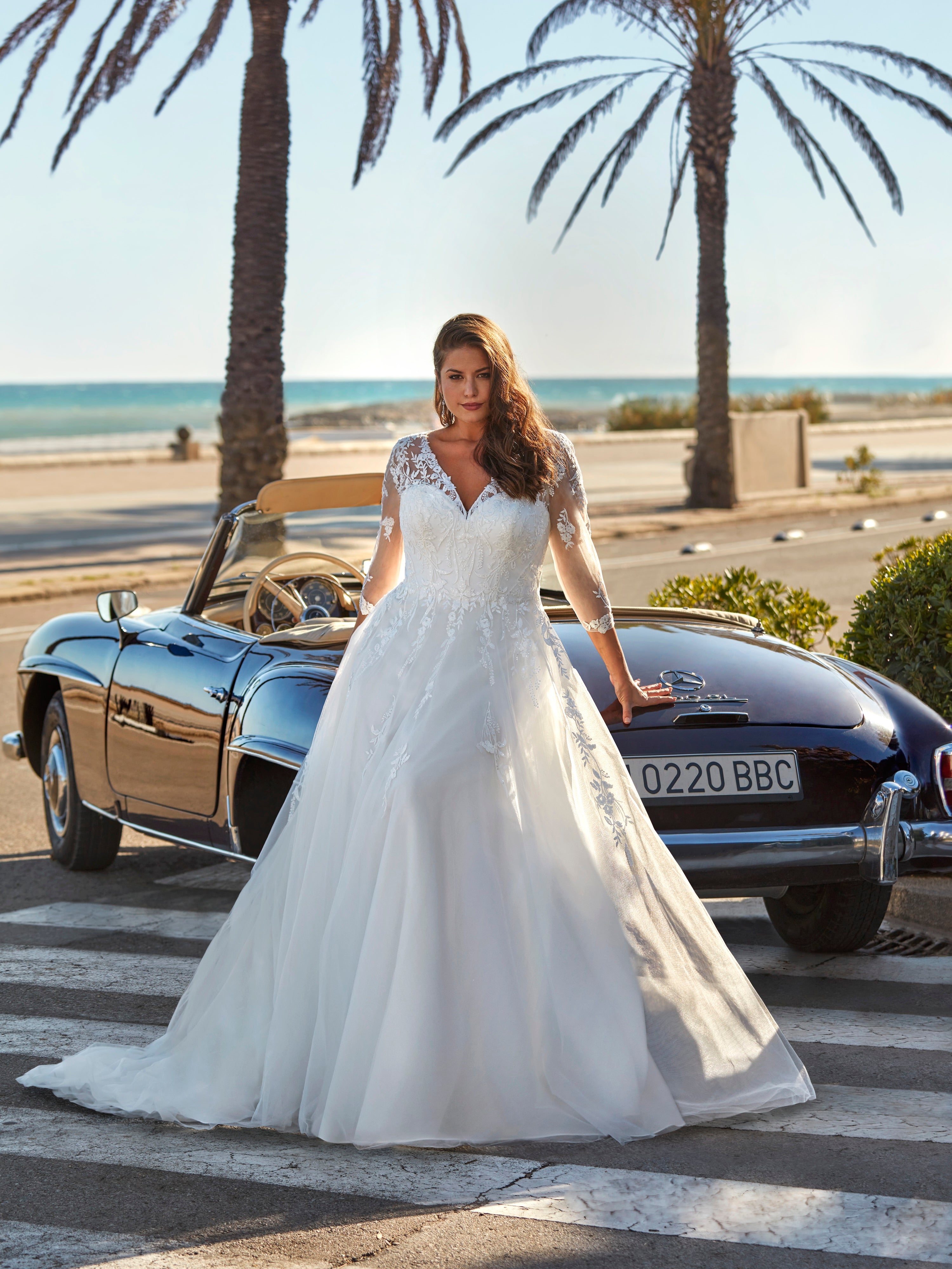 Woman in a white wedding dress standing next to a vintage car on a beach road.
