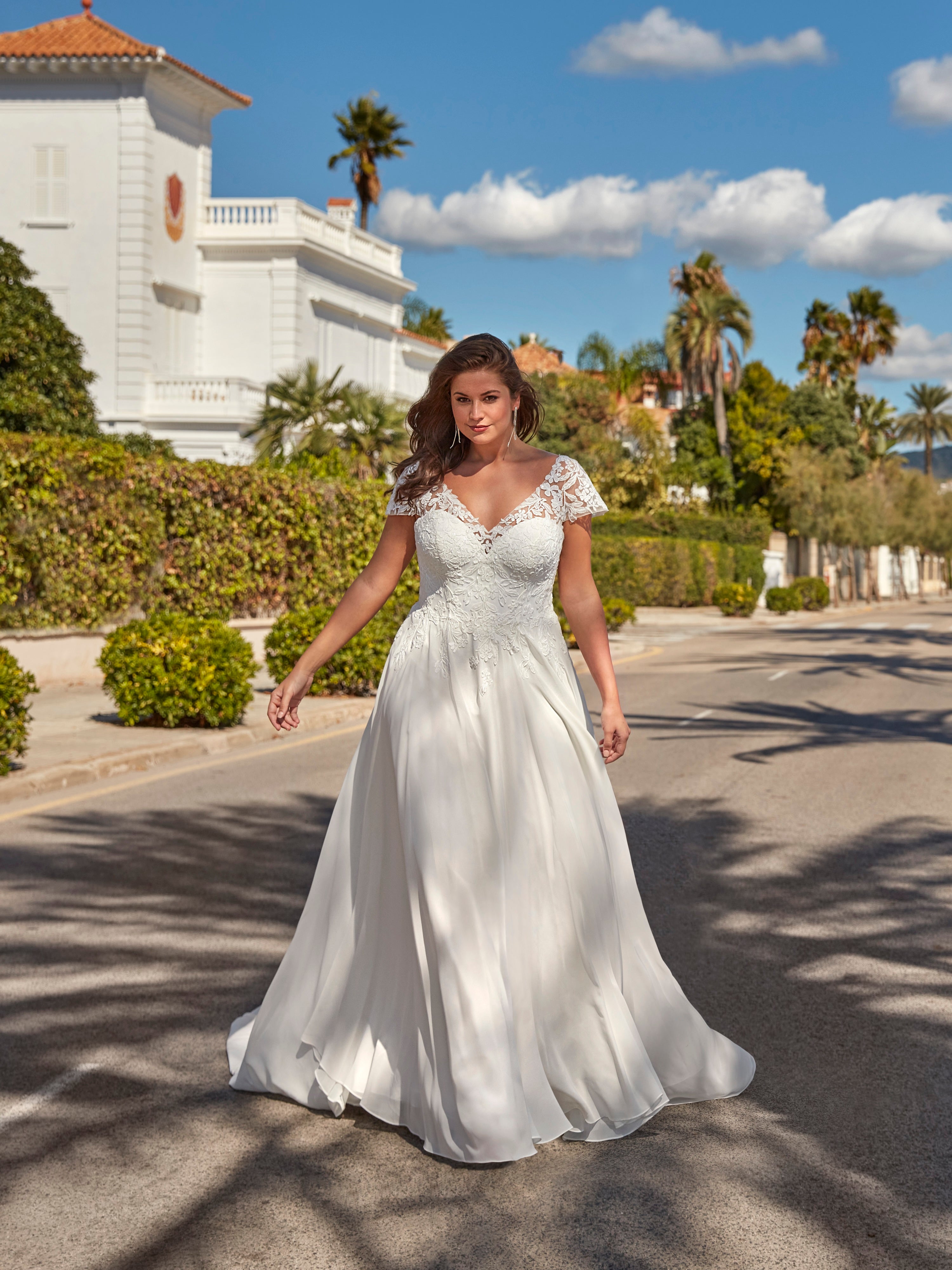 Woman in a white wedding dress standing on a street with palm trees and a building in the background.