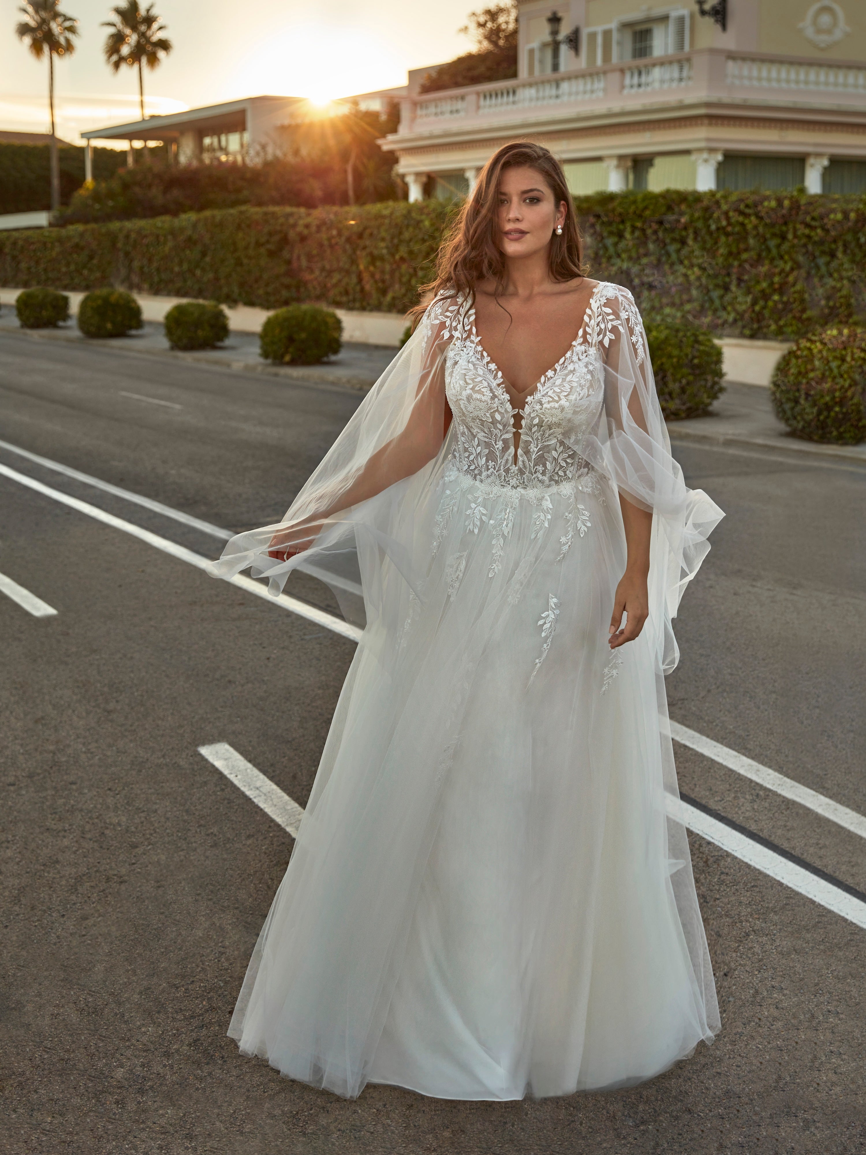 Woman in a white wedding dress with a cape standing on a street.