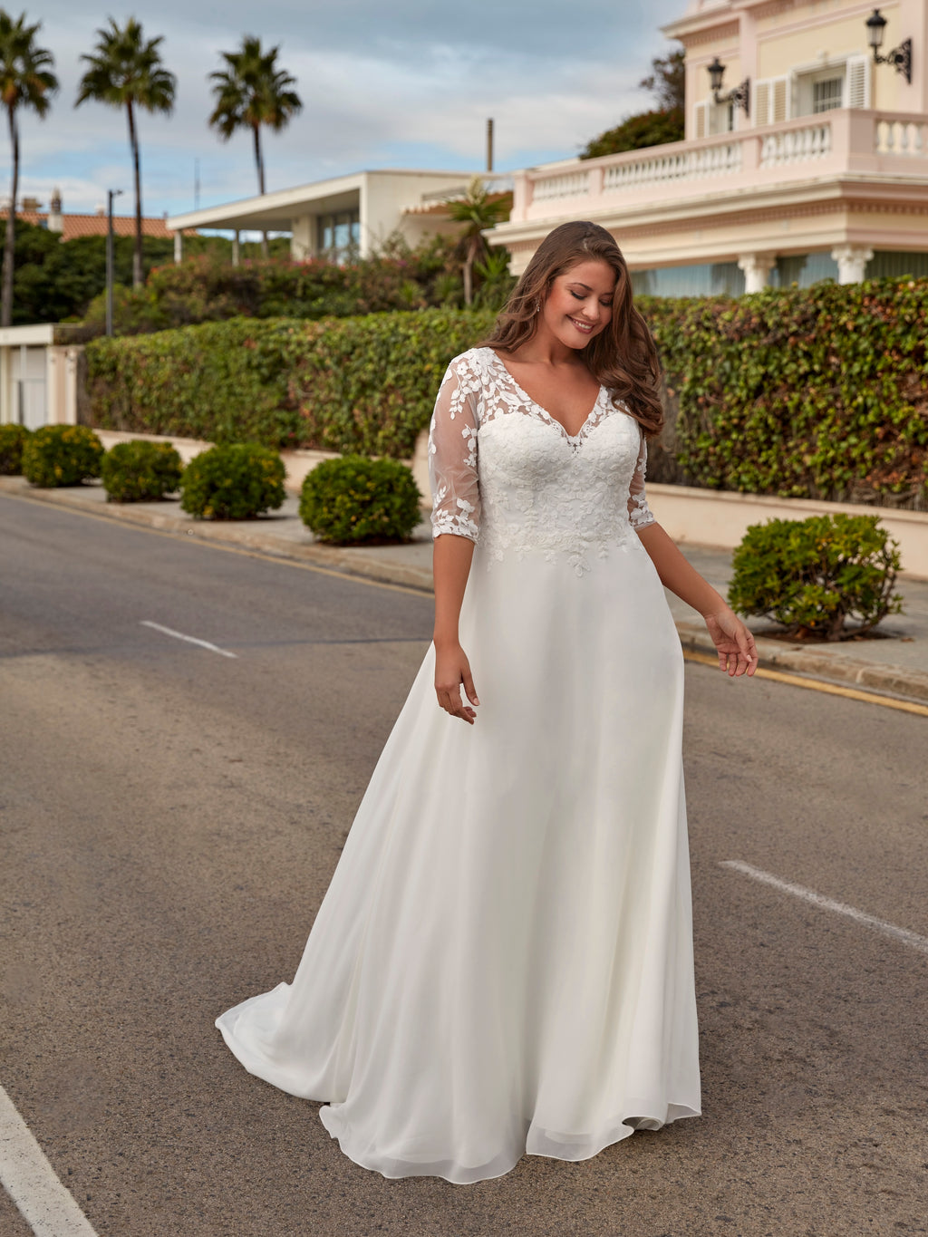 Woman in a white lace wedding dress standing on a street with palm trees and a building in the background.