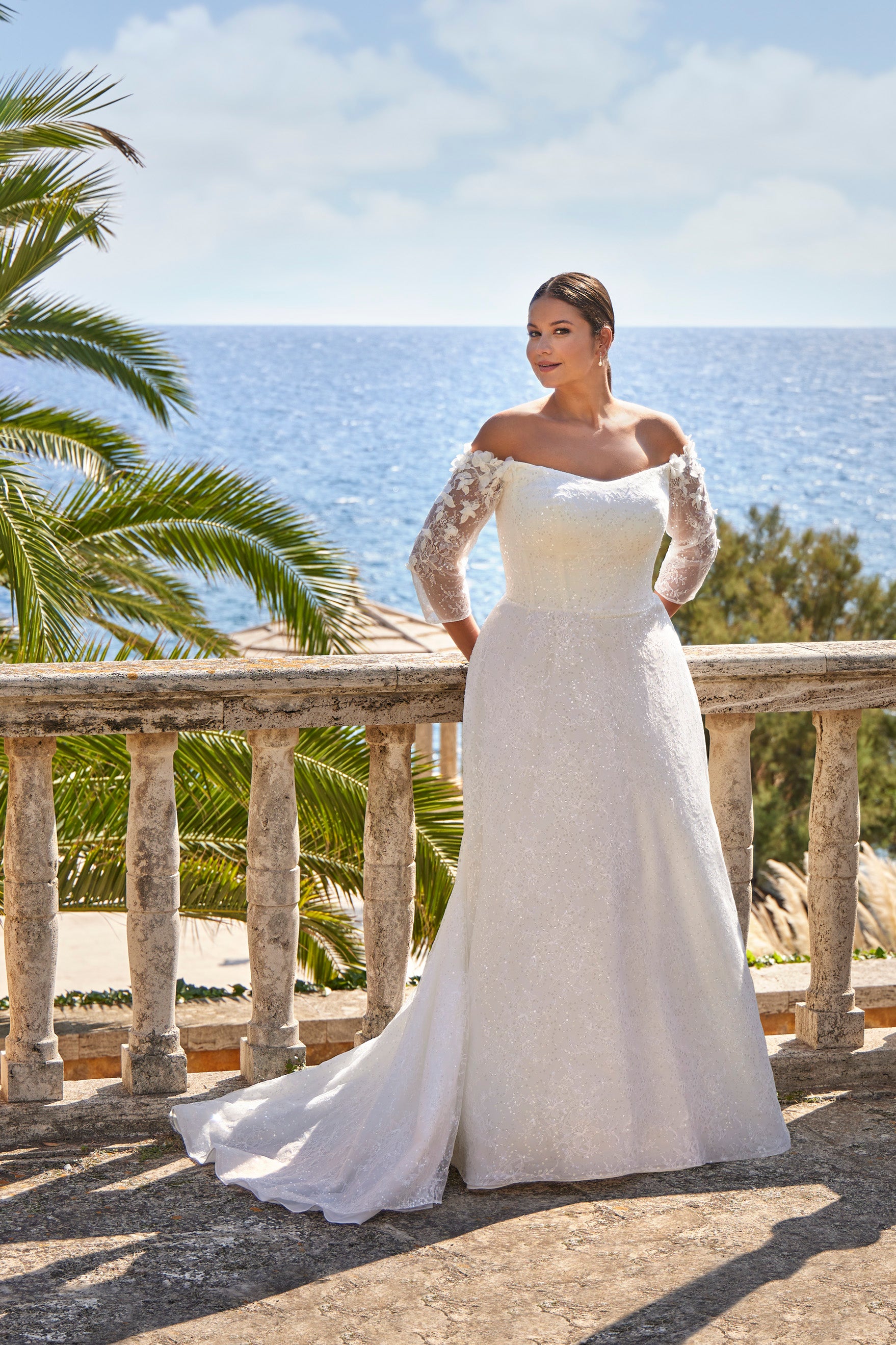 Woman in a white wedding dress with lace sleeves standing on a balcony with ocean view