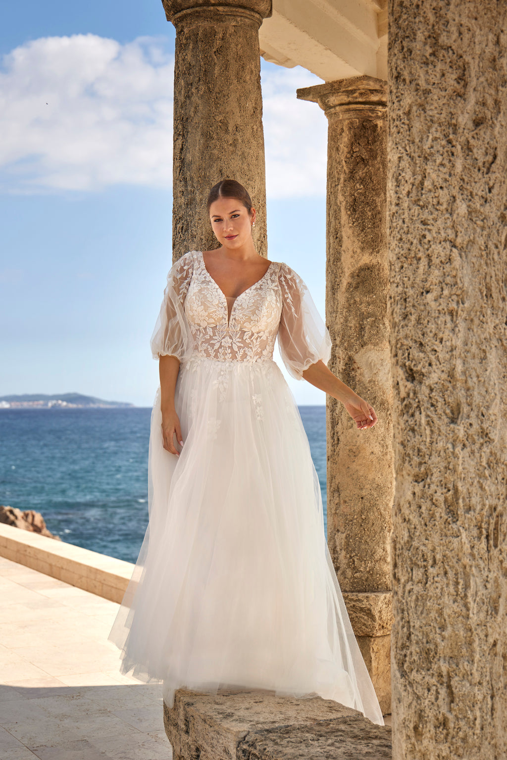 Woman in a white lace dress standing by stone columns with ocean and sky in the background