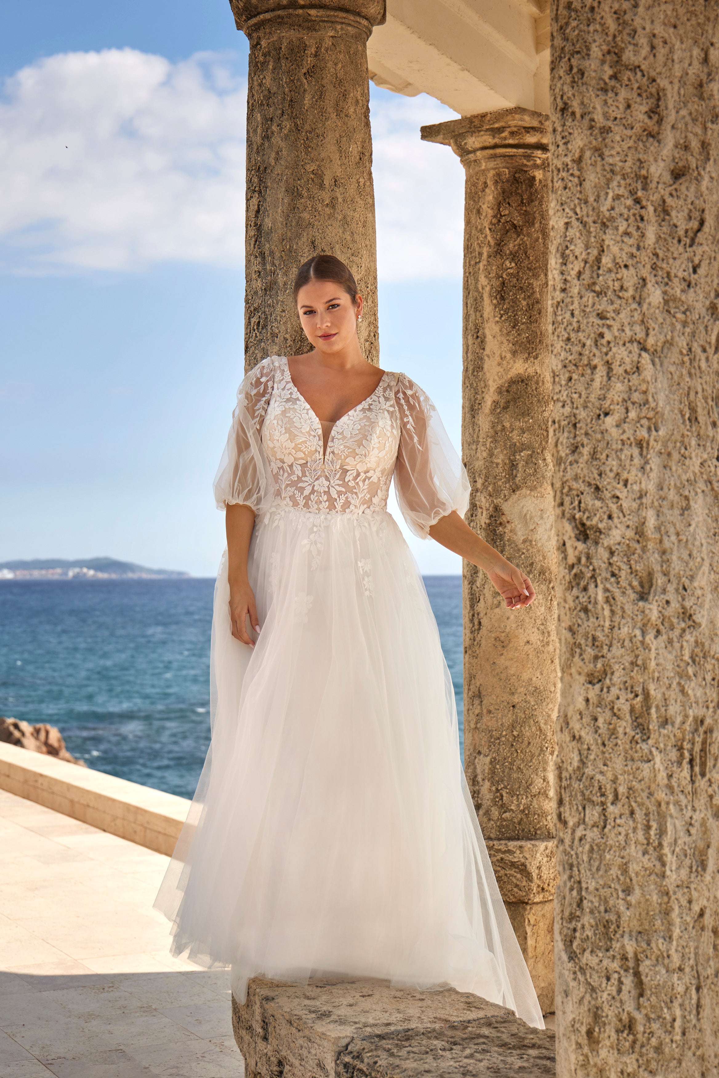 Woman in a white lace dress standing by stone columns with ocean and sky in the background