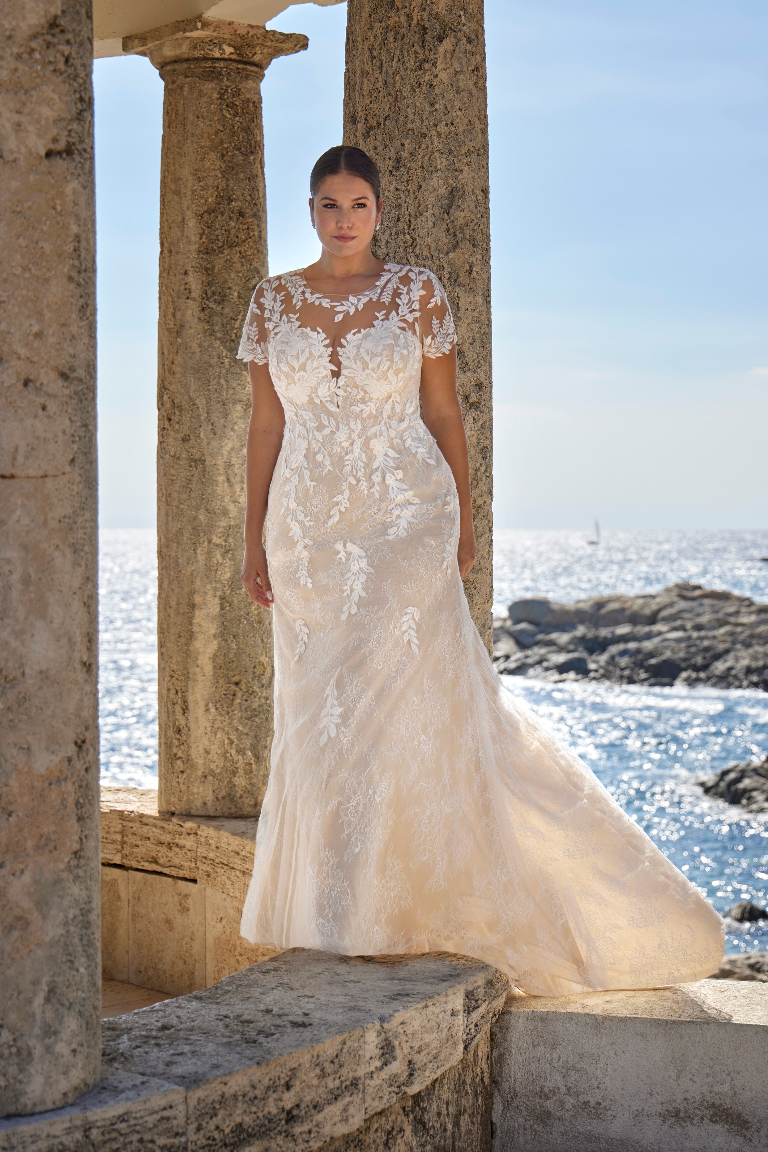 Woman in a white lace wedding dress standing in front of stone columns with a coastal background.