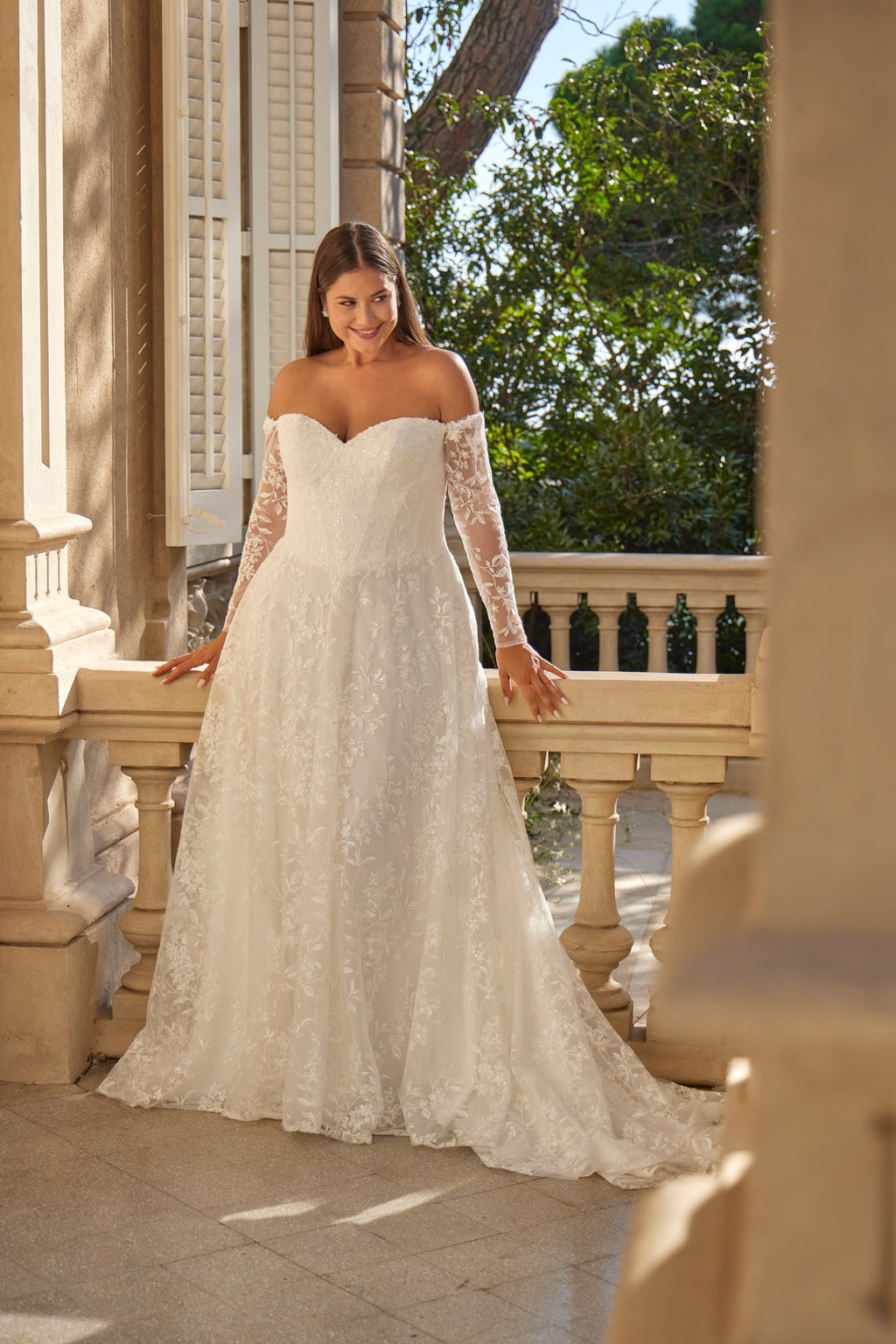 Woman in a white lace wedding dress standing on a balcony with greenery in the background