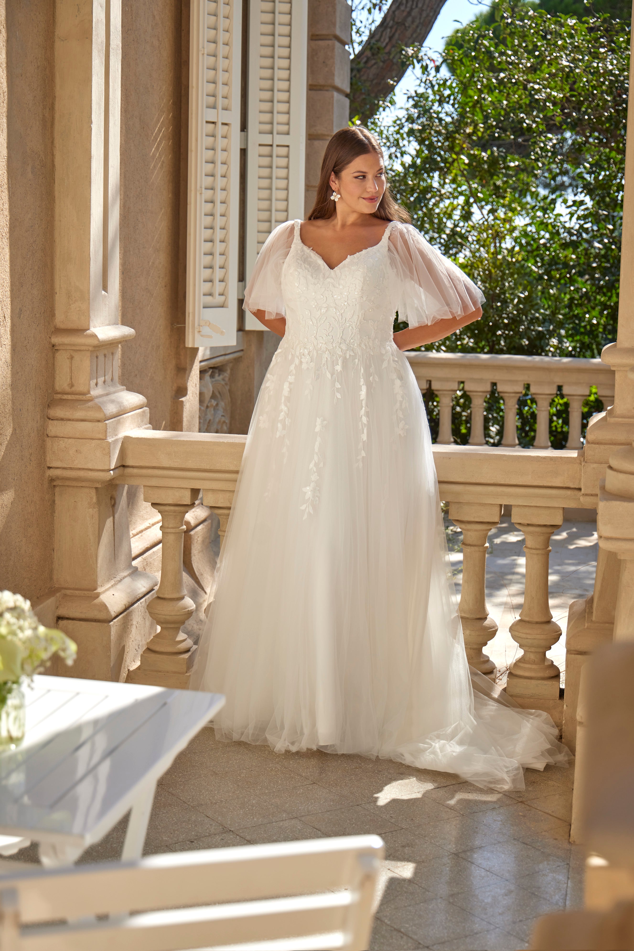 Woman in a white wedding dress standing on a balcony with greenery in the background