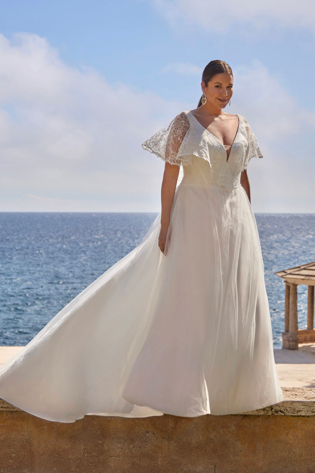 Woman in a white wedding dress standing by the ocean with a clear blue sky.