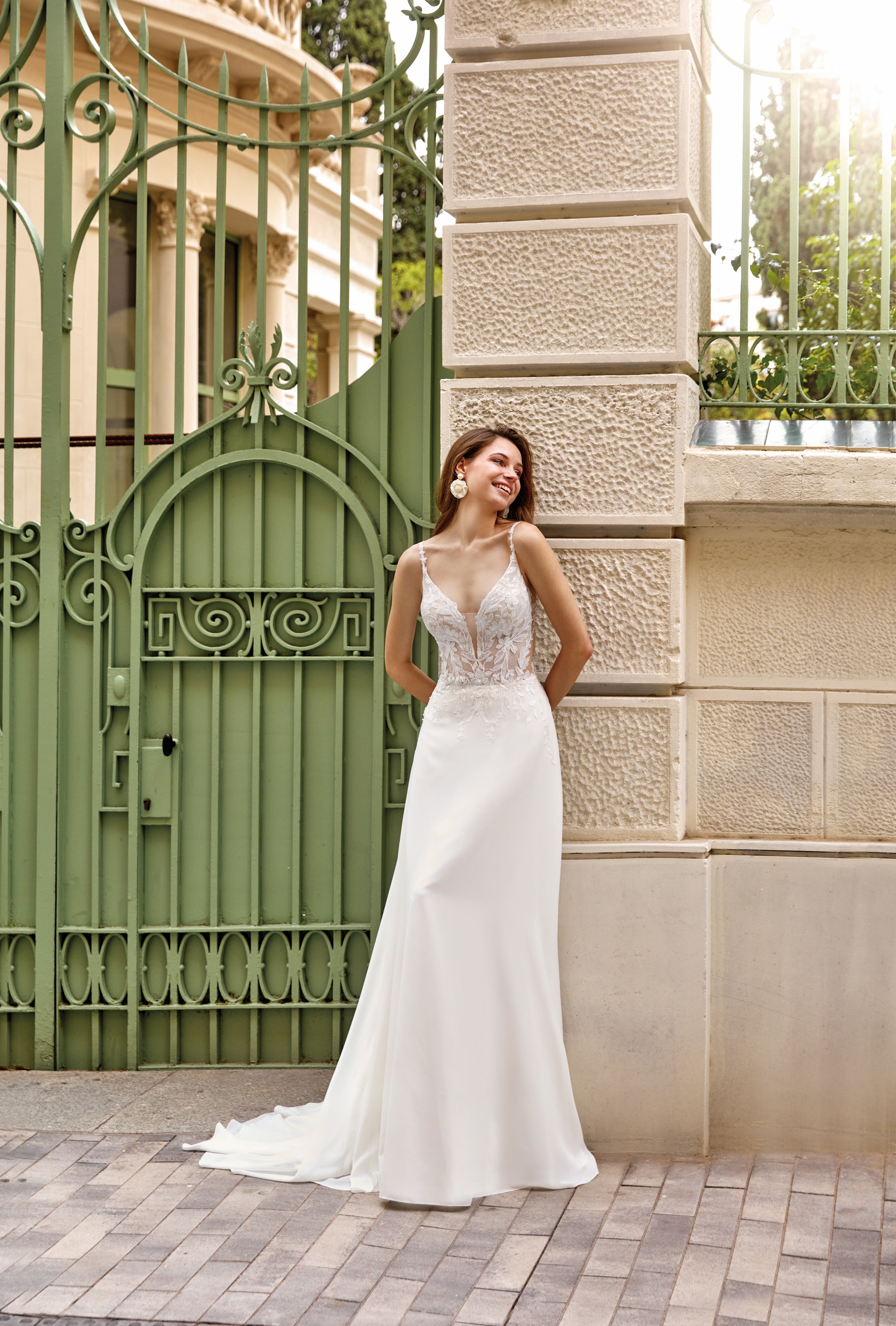 Woman in a white wedding dress standing in front of a decorative green gate and stone building.