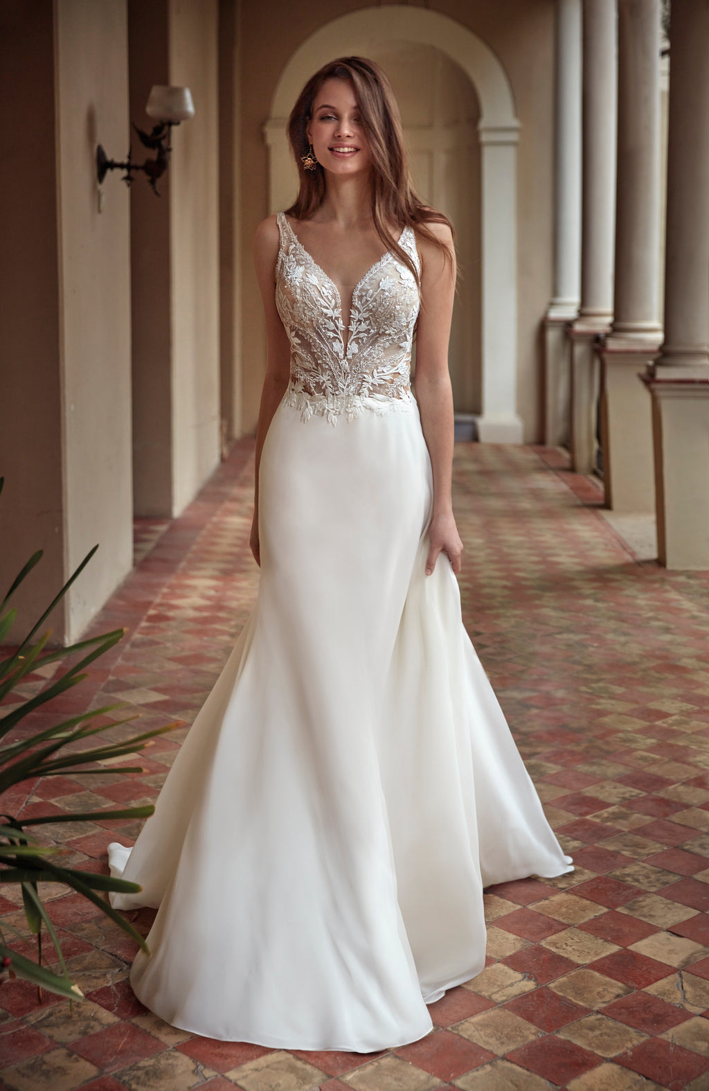 Woman in a white wedding dress with intricate detailing standing in a sunlit hallway.