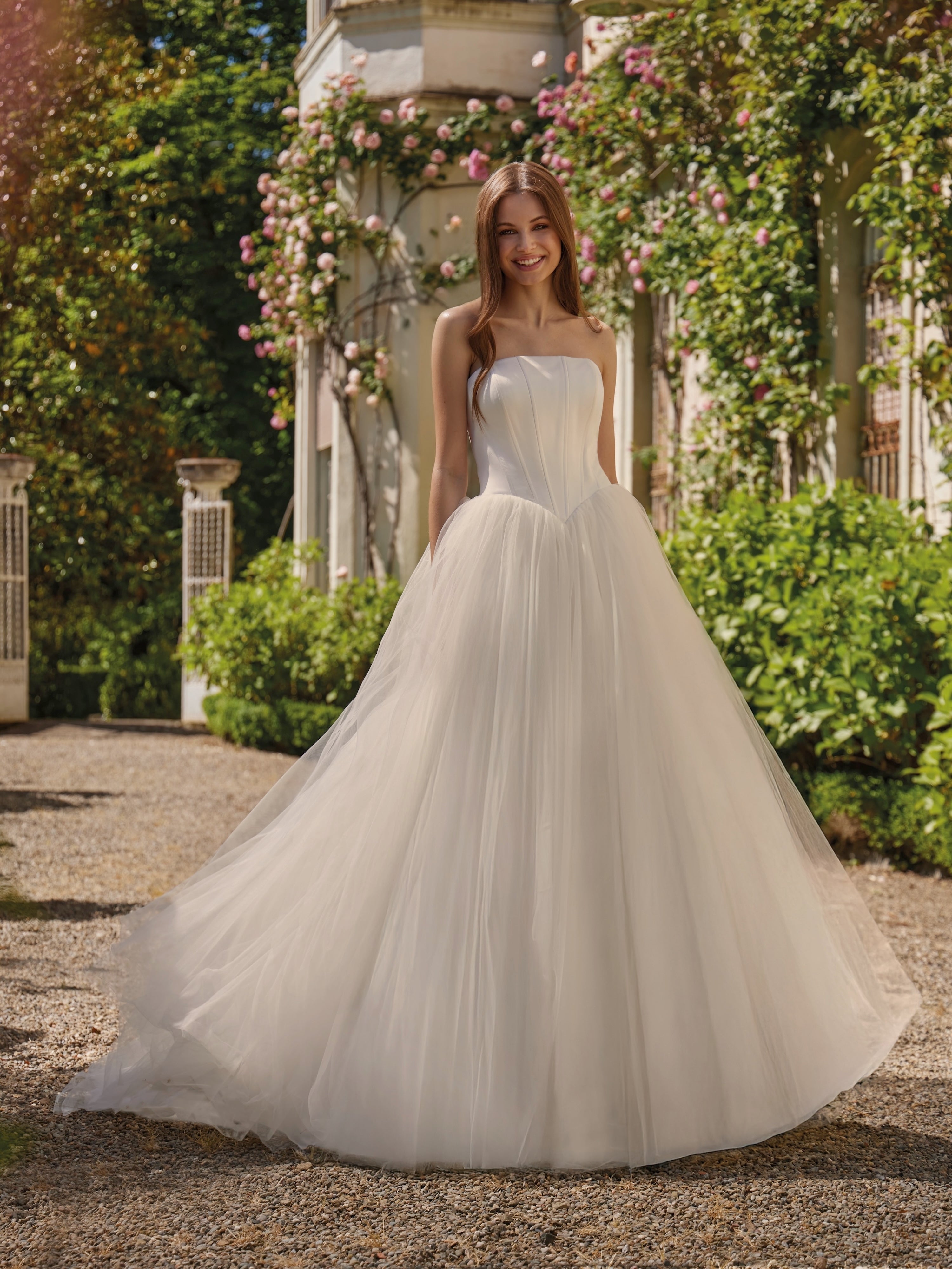 Woman in a white strapless wedding dress standing outdoors with greenery and flowers in the background