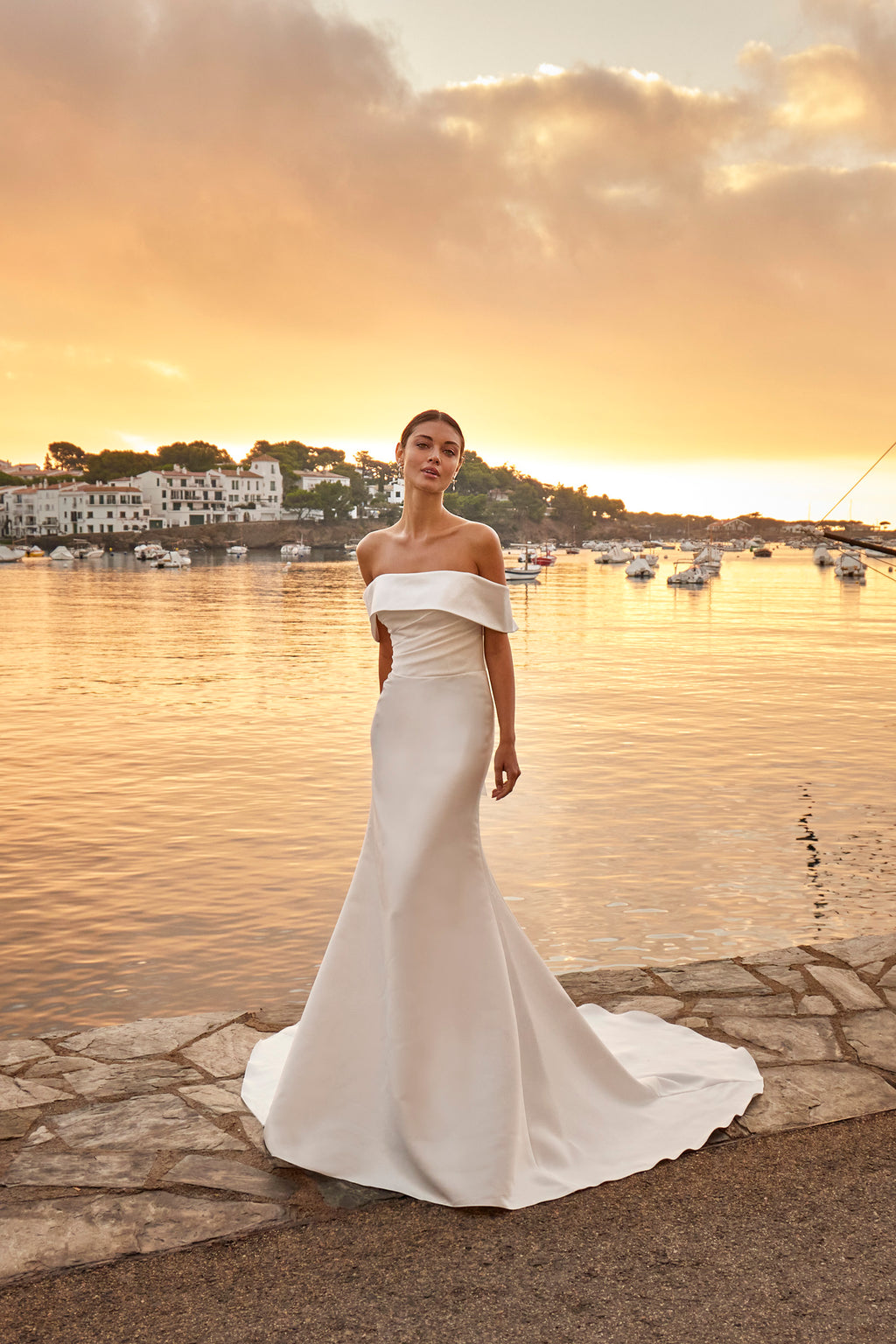 Woman in a white off-shoulder gown standing by a waterfront at sunset.