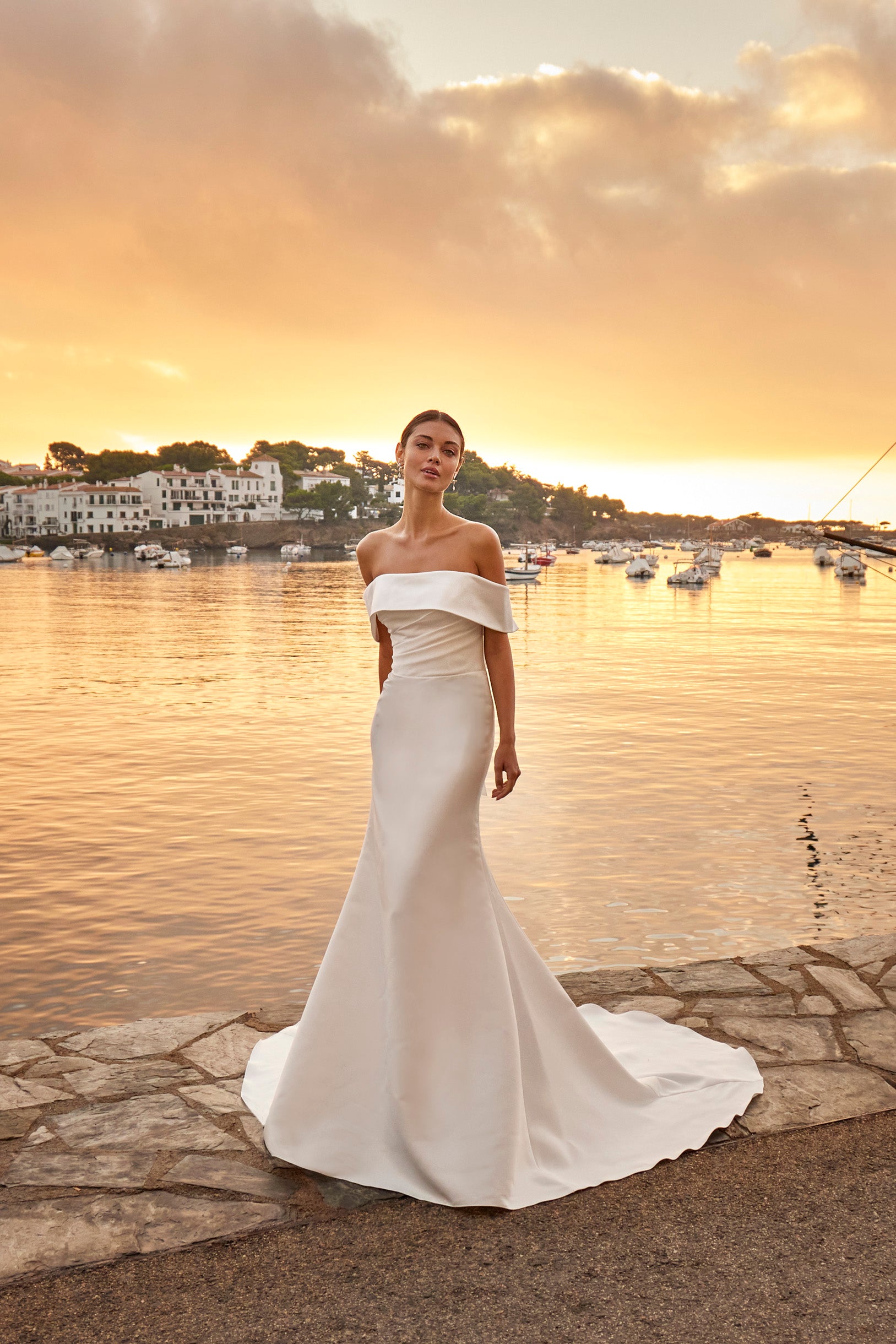 Woman in a white off-shoulder gown standing by a waterfront at sunset.