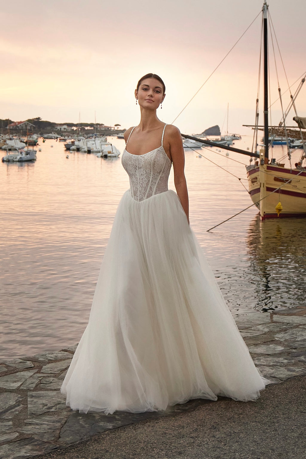 Woman in a white wedding dress standing by a waterfront with boats and sunset.
