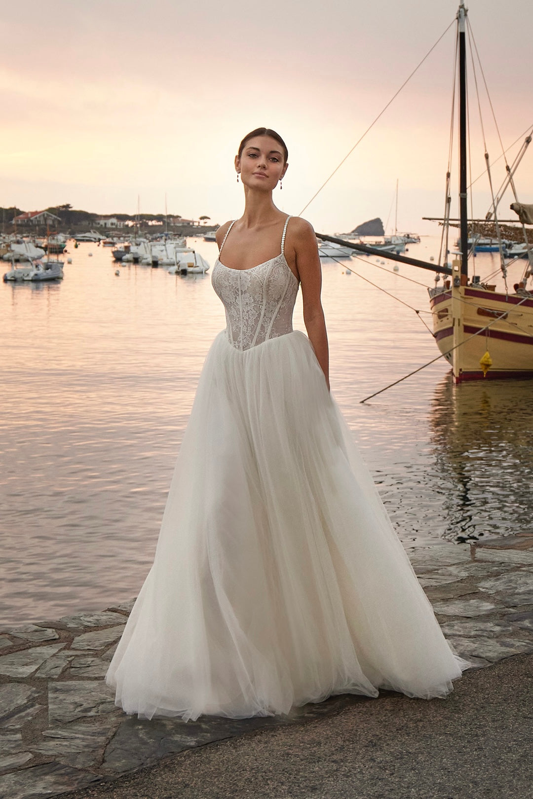 Woman in a white wedding dress standing by a waterfront with boats and sunset.