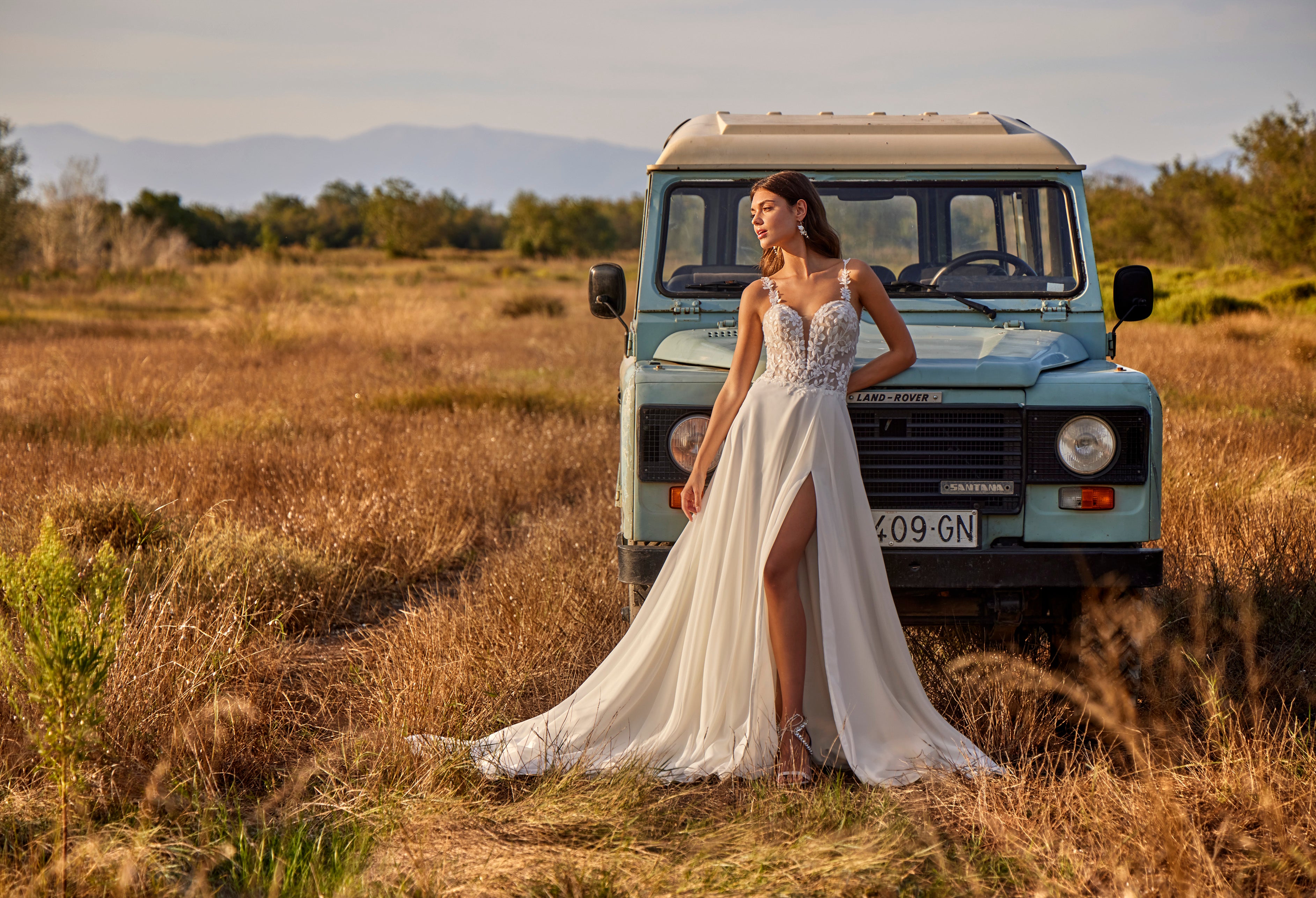Woman in a white dress standing next to a vintage Land Rover in a field with mountains in the background