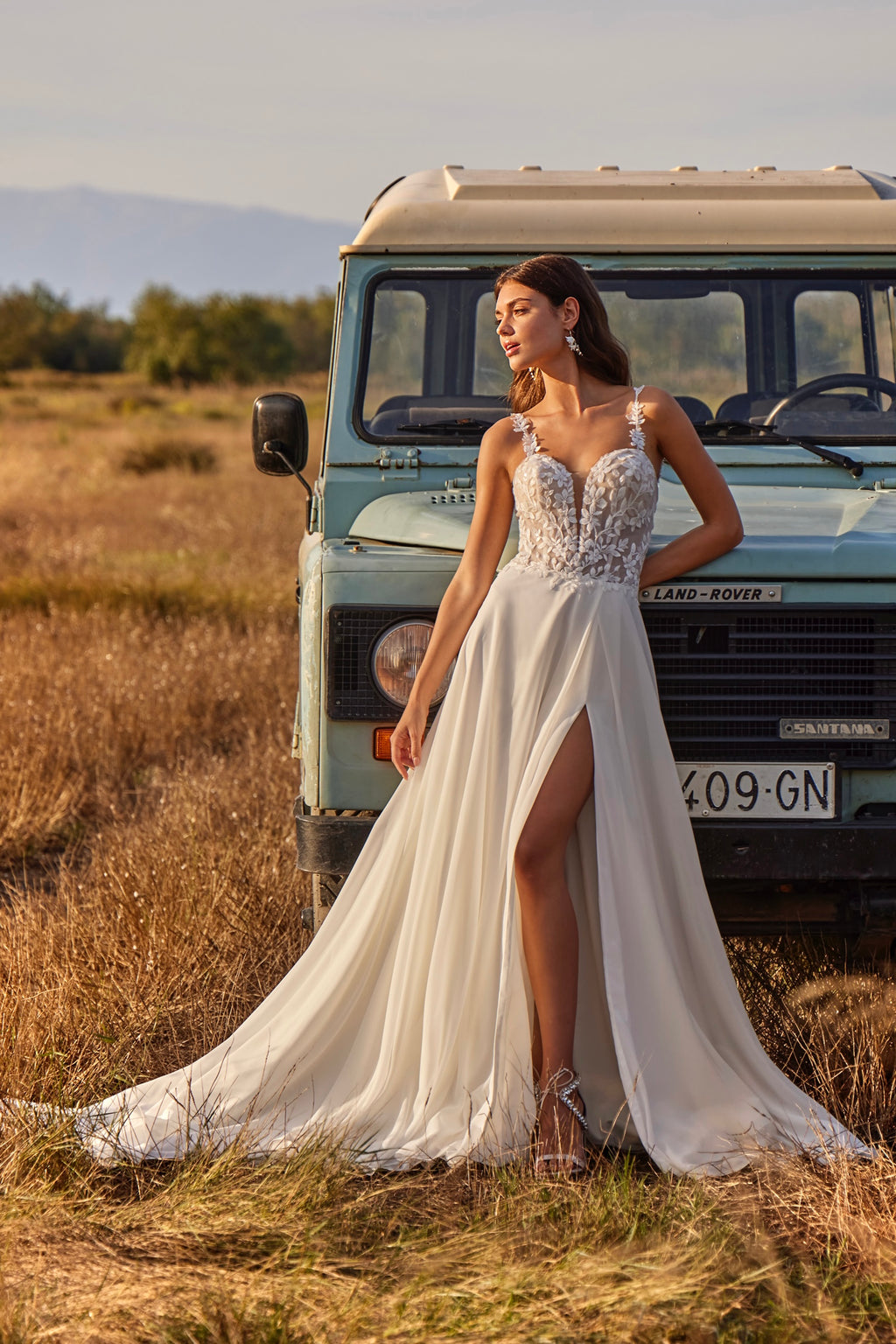 Woman in a white wedding dress standing in front of a vintage Land Rover in a field.
