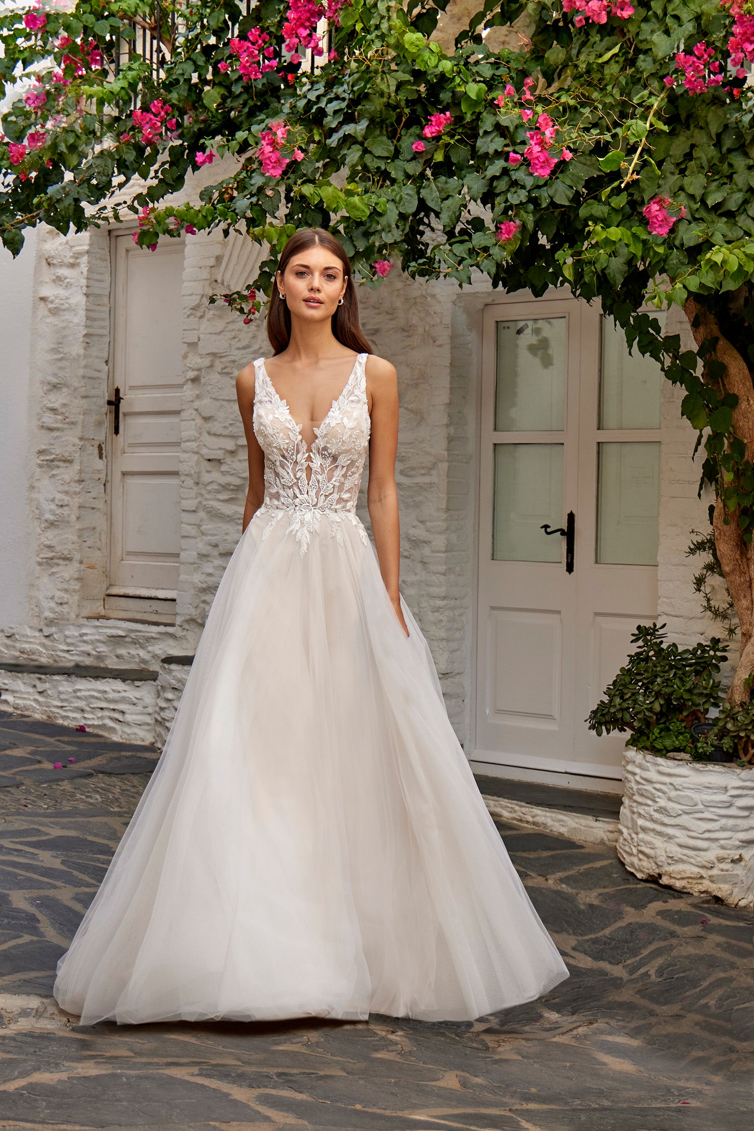 Woman in a white wedding dress standing in front of a stone building with pink flowers.