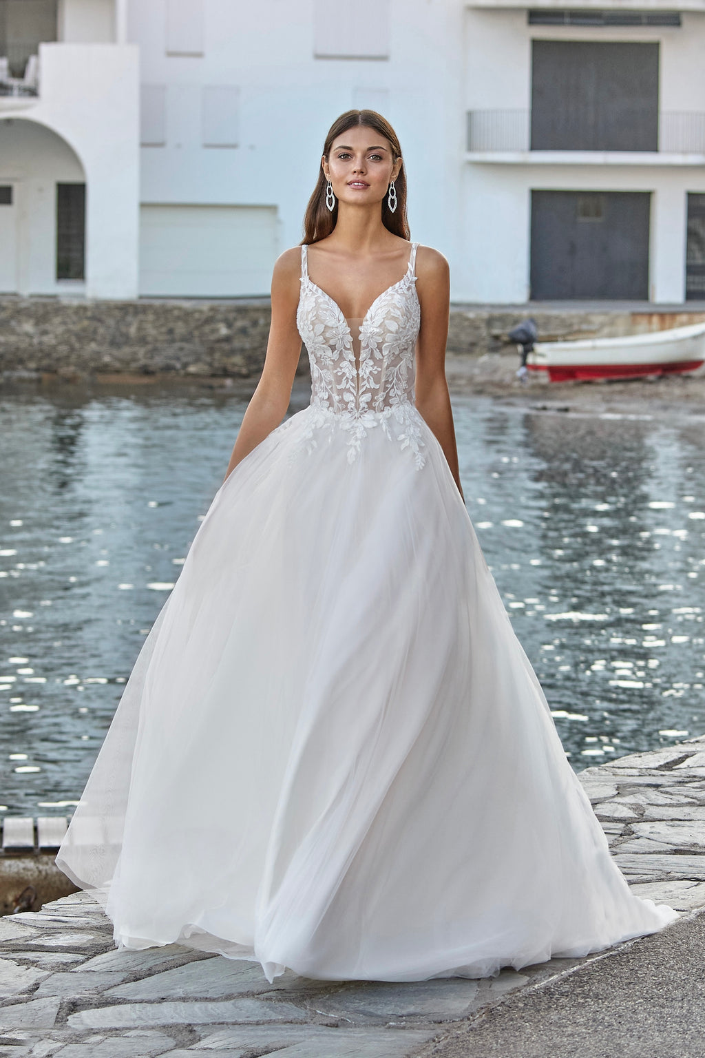 Woman in a white wedding dress standing by a waterfront with a building and boat in the background.