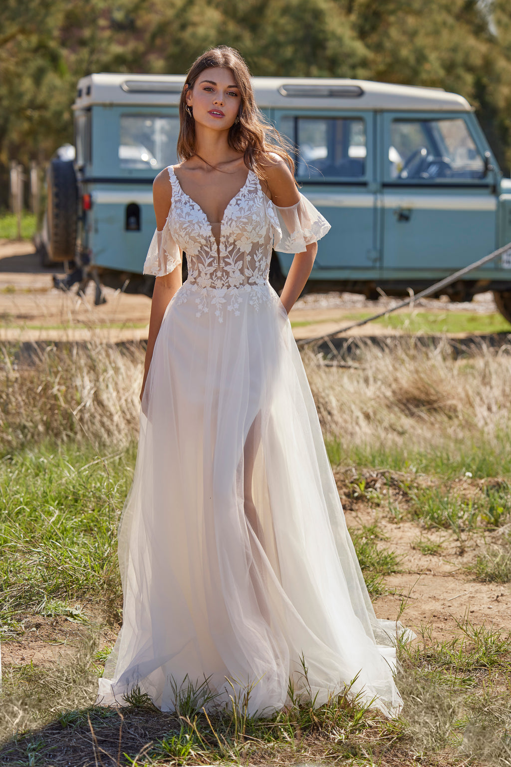 Woman in a white wedding dress standing in a natural setting with a vintage van in the background.