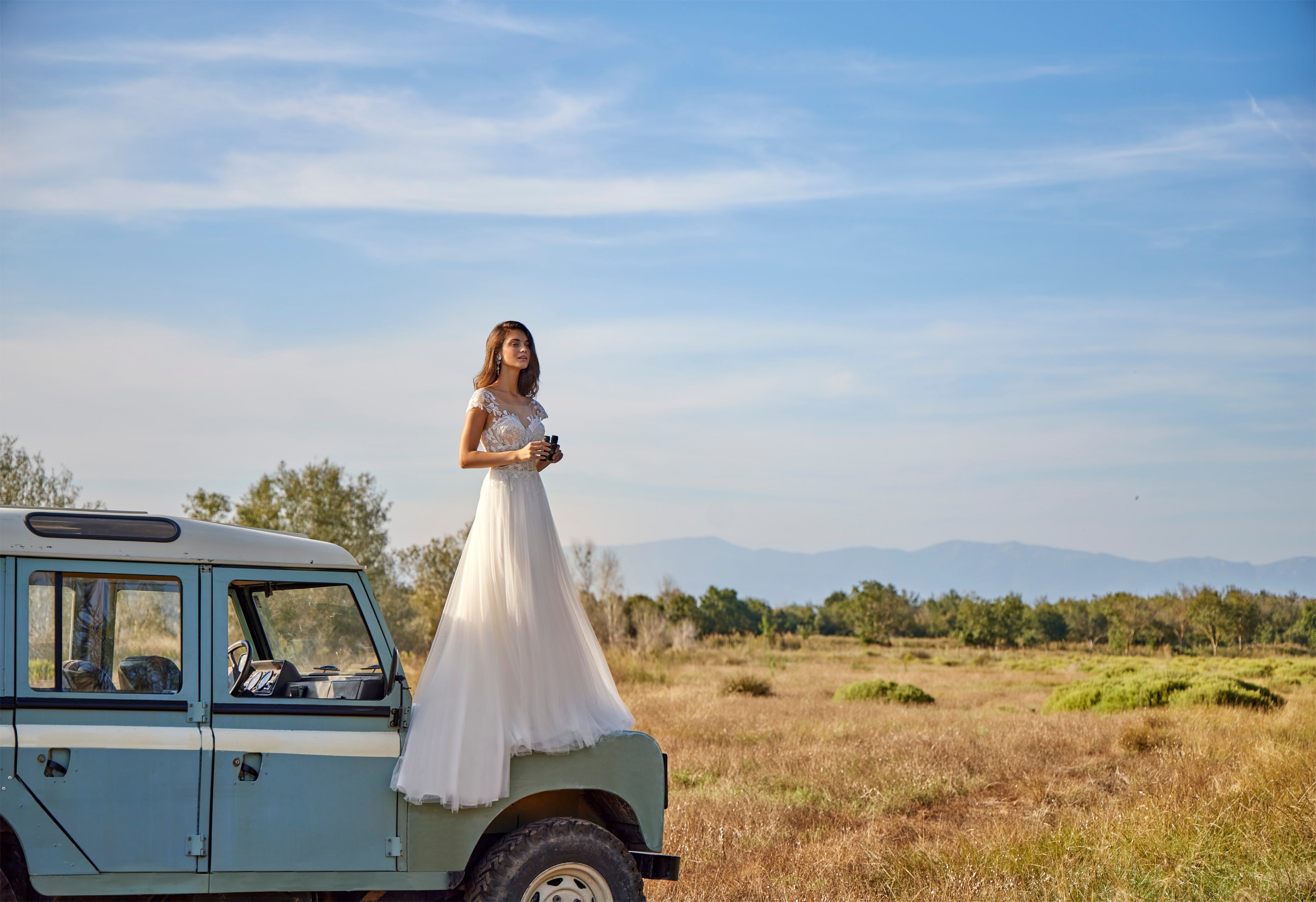 Woman in a white dress standing on an old-fashioned car in a field with a blue sky.