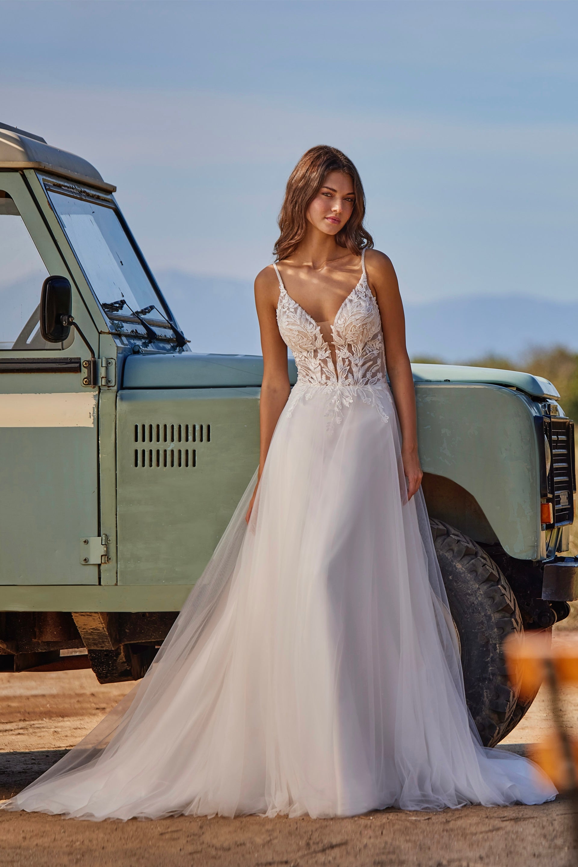 Woman in a white wedding dress standing next to a vintage vehicle with a scenic background