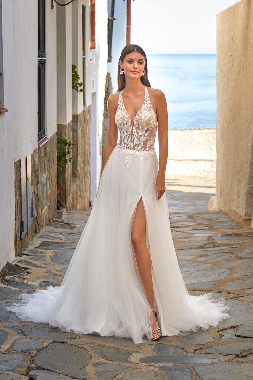 Woman in a white wedding dress with a high slit standing on a stone path near a coastal building.