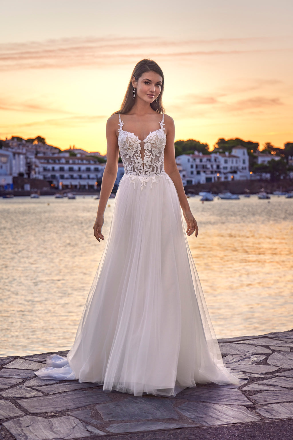 Woman in a white wedding dress standing by a waterfront at sunset.